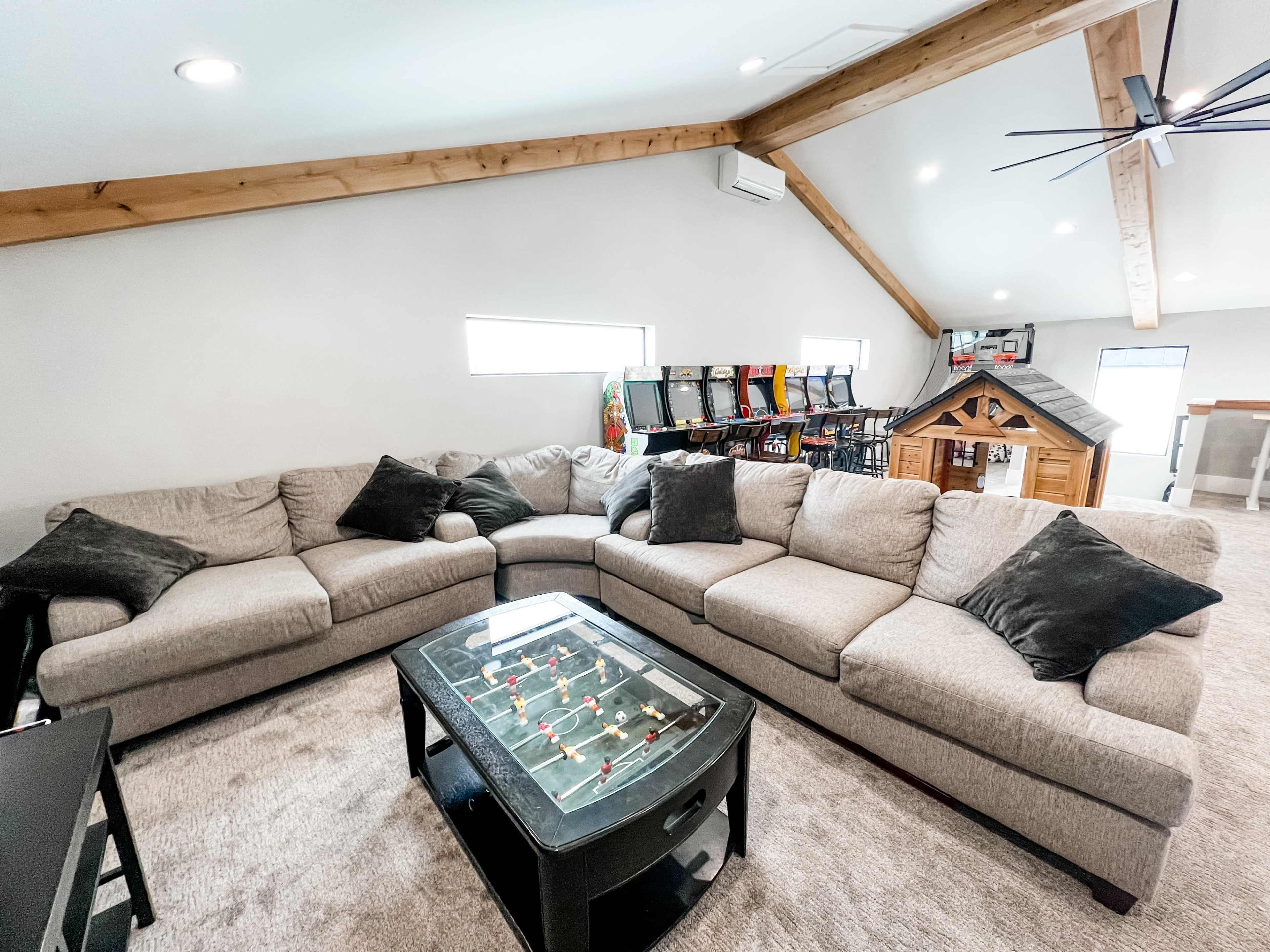 A cozy loft living area featuring a large sectional sofa, a glass coffee table with a foosball setup, and a small playhouse in the corner.