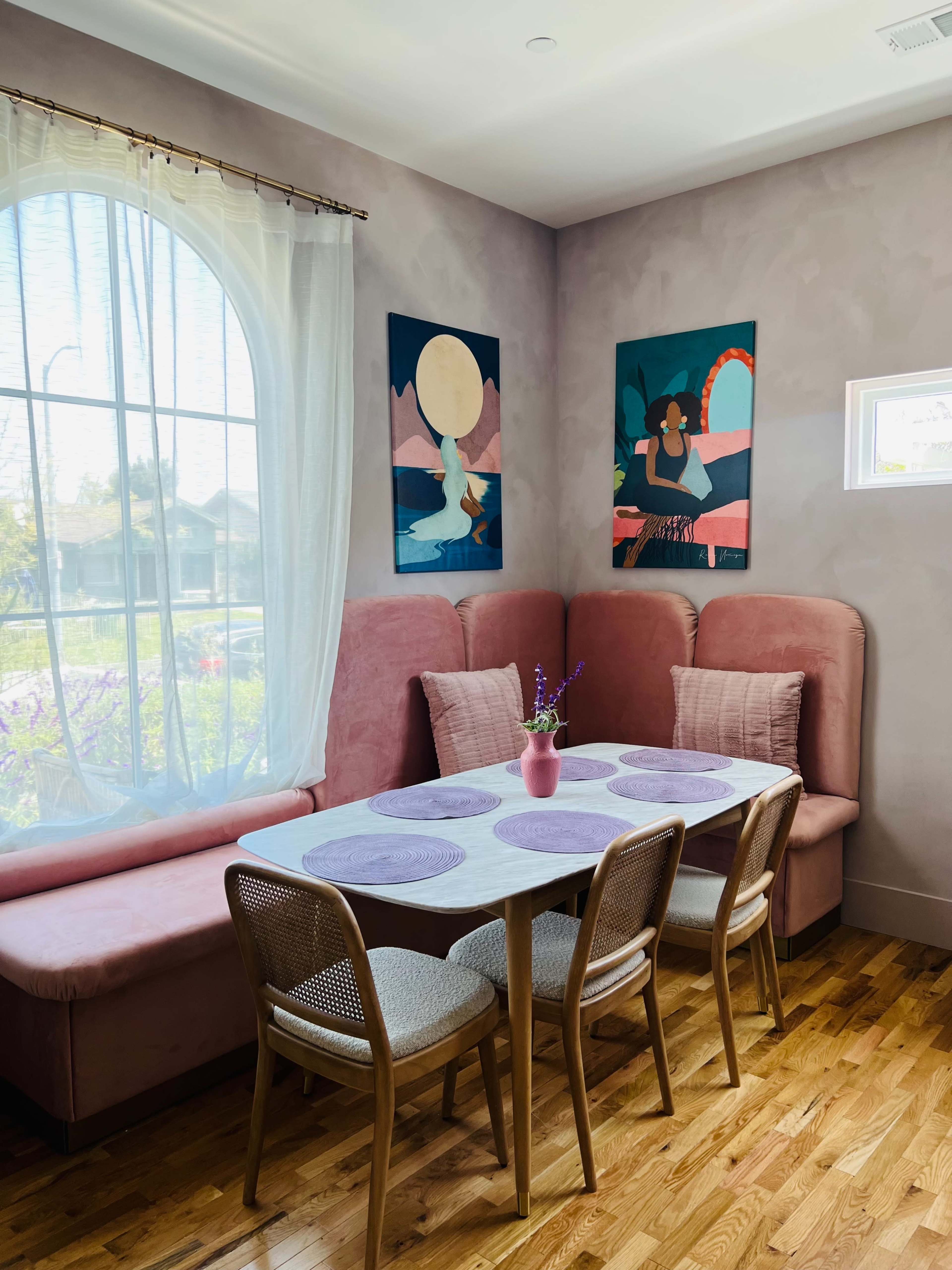 A dining area features a white table with purple placemats surrounded by four woven chairs, set against a backdrop of pink upholstered seating and two colorful art pieces on the wall.