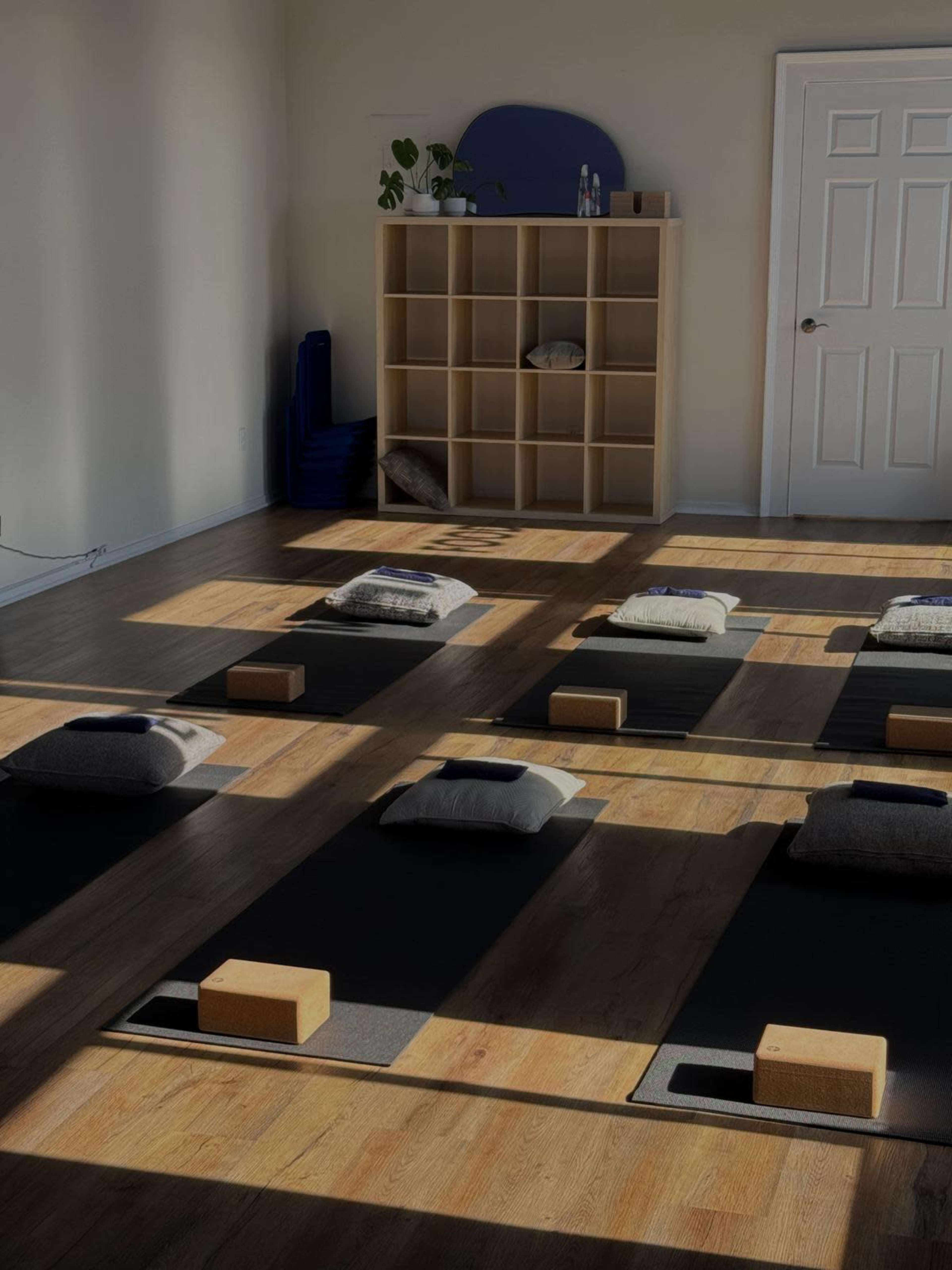 A spacious yoga studio with several mats arranged on the floor, each equipped with blocks and cushions, illuminated by sunlight streaming through the windows.
