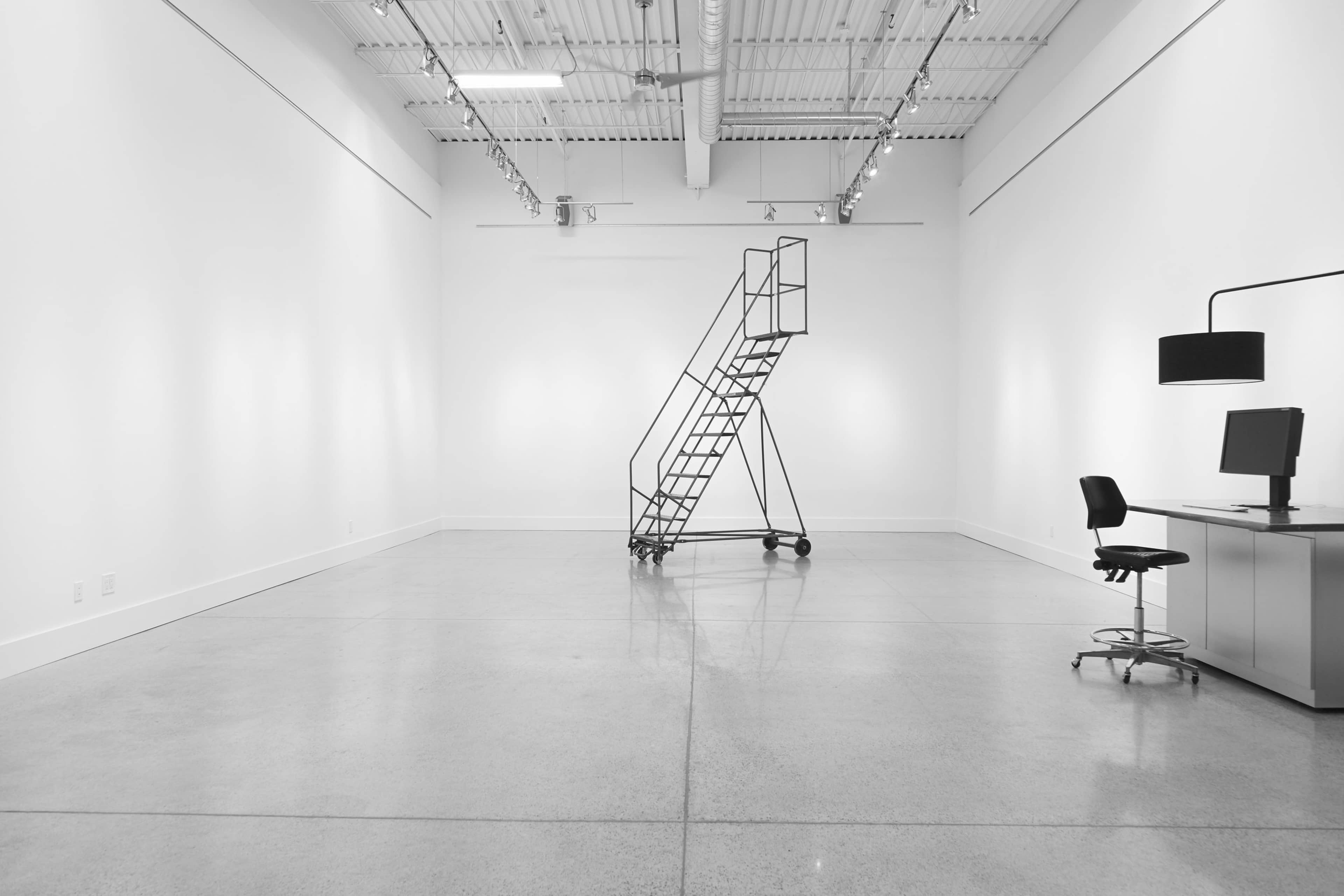 A spacious, minimalistic room with a metal ladder, a desk with a chair, and a computer setup against a stark white wall.
