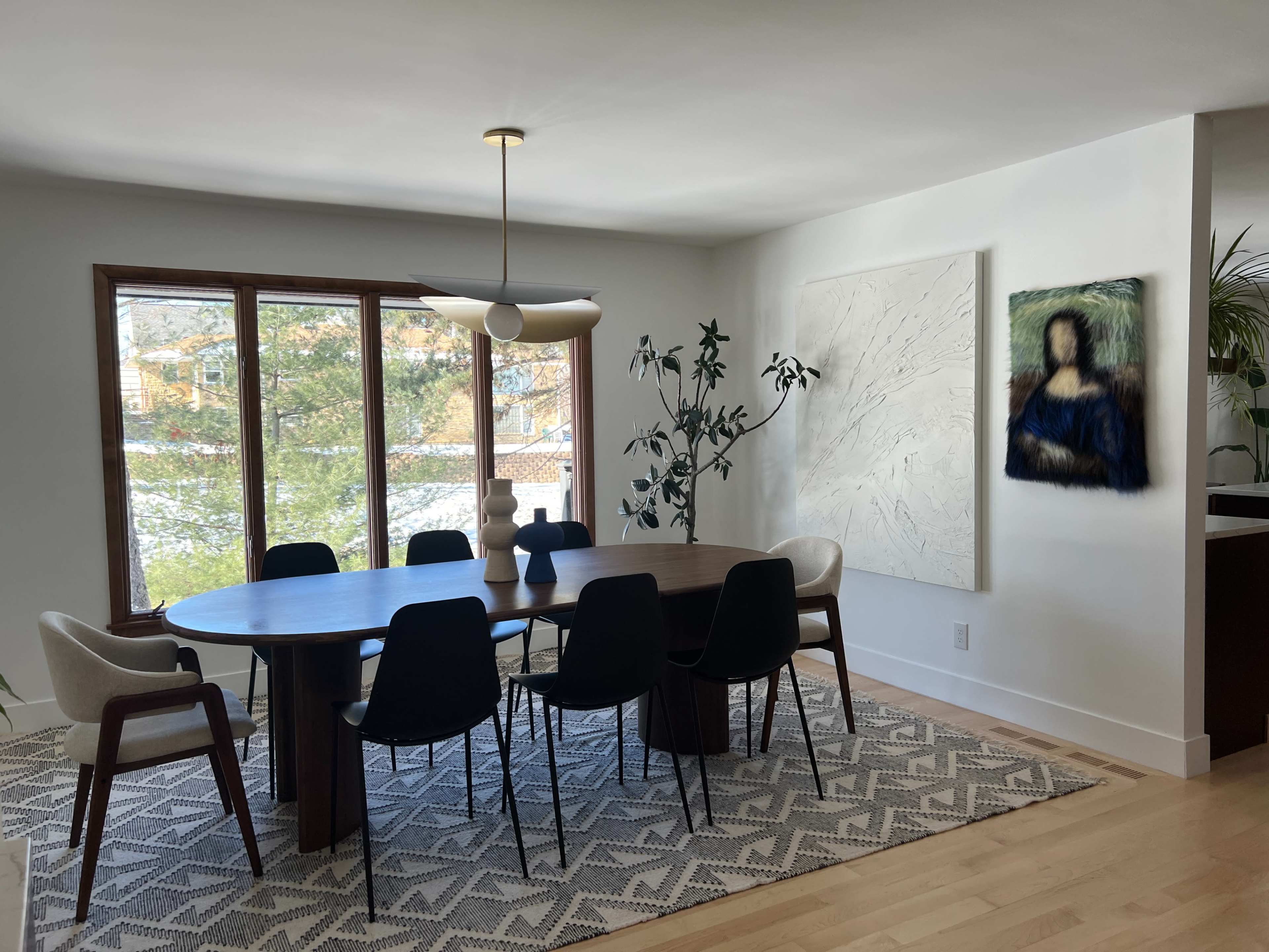 A modern dining area features a large round table surrounded by black chairs, with a prominent artwork on the wall and large windows letting in natural light.