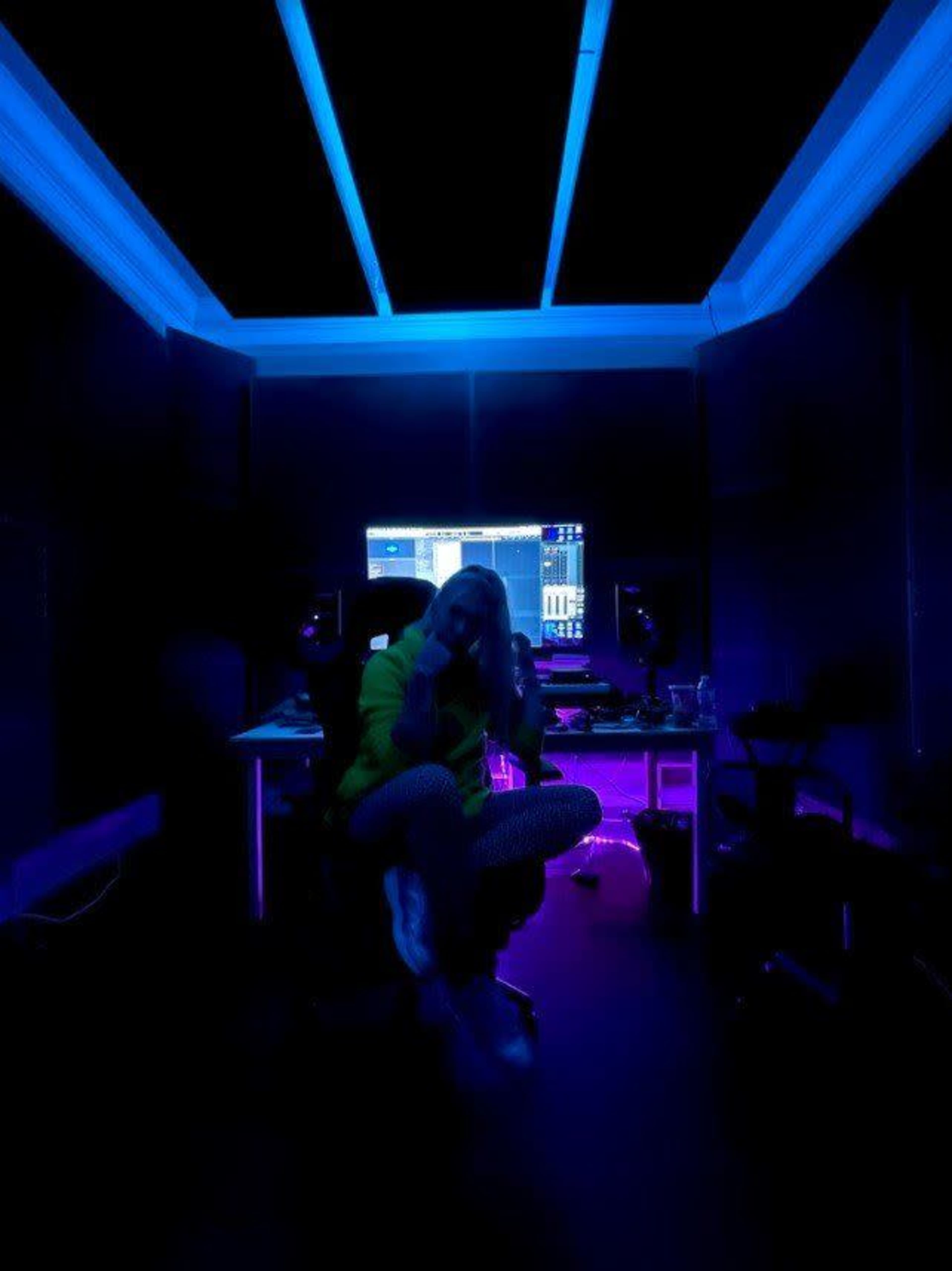A person sits cross-legged in a dimly lit recording studio, with blue and purple lighting illuminating the space and a computer screen displaying audio software in the background.