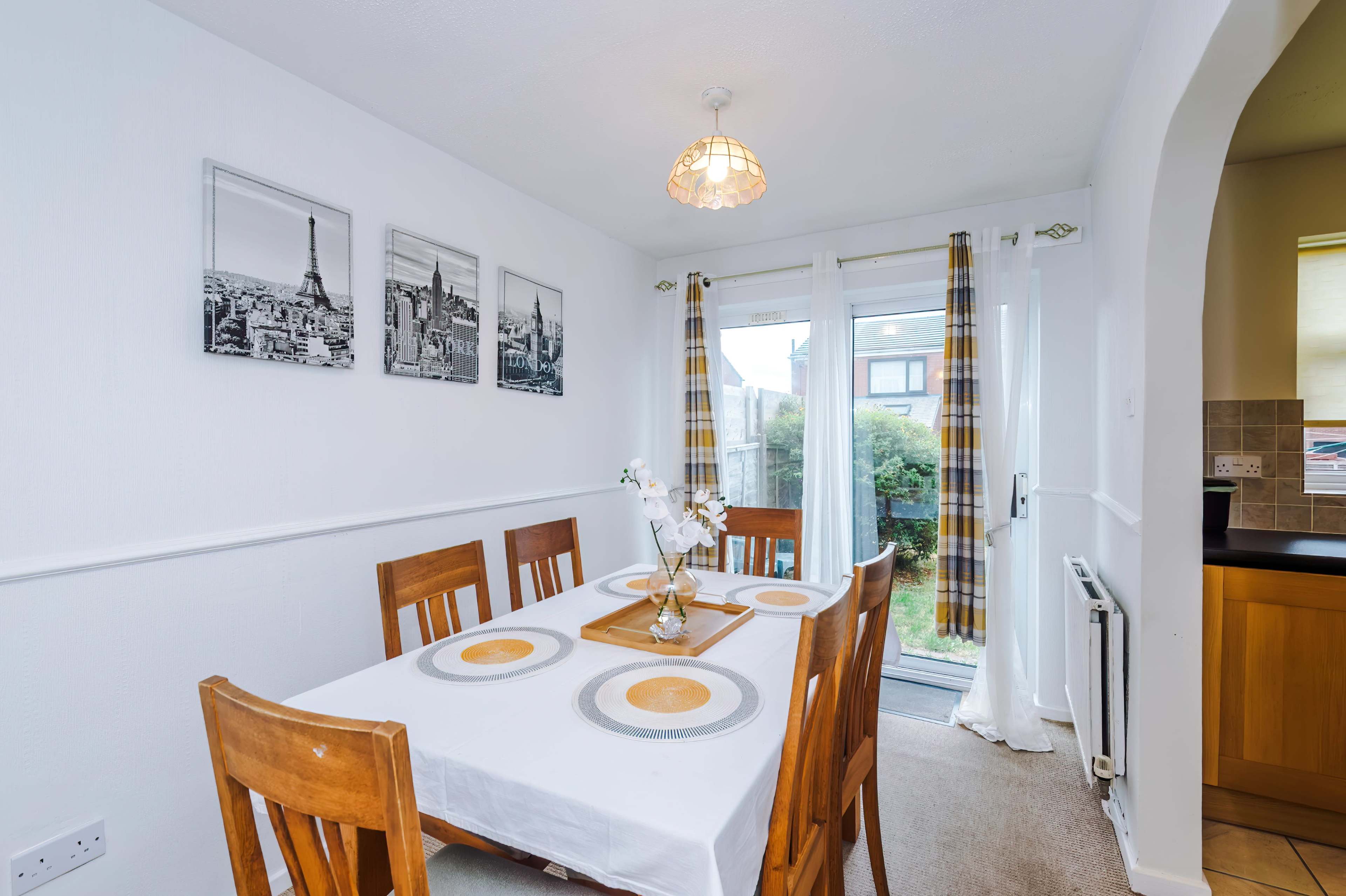 The image shows a dining area with a table surrounded by wooden chairs, featuring a white tablecloth and wall art depicting cityscapes, along with a glass door leading to an outdoor space.