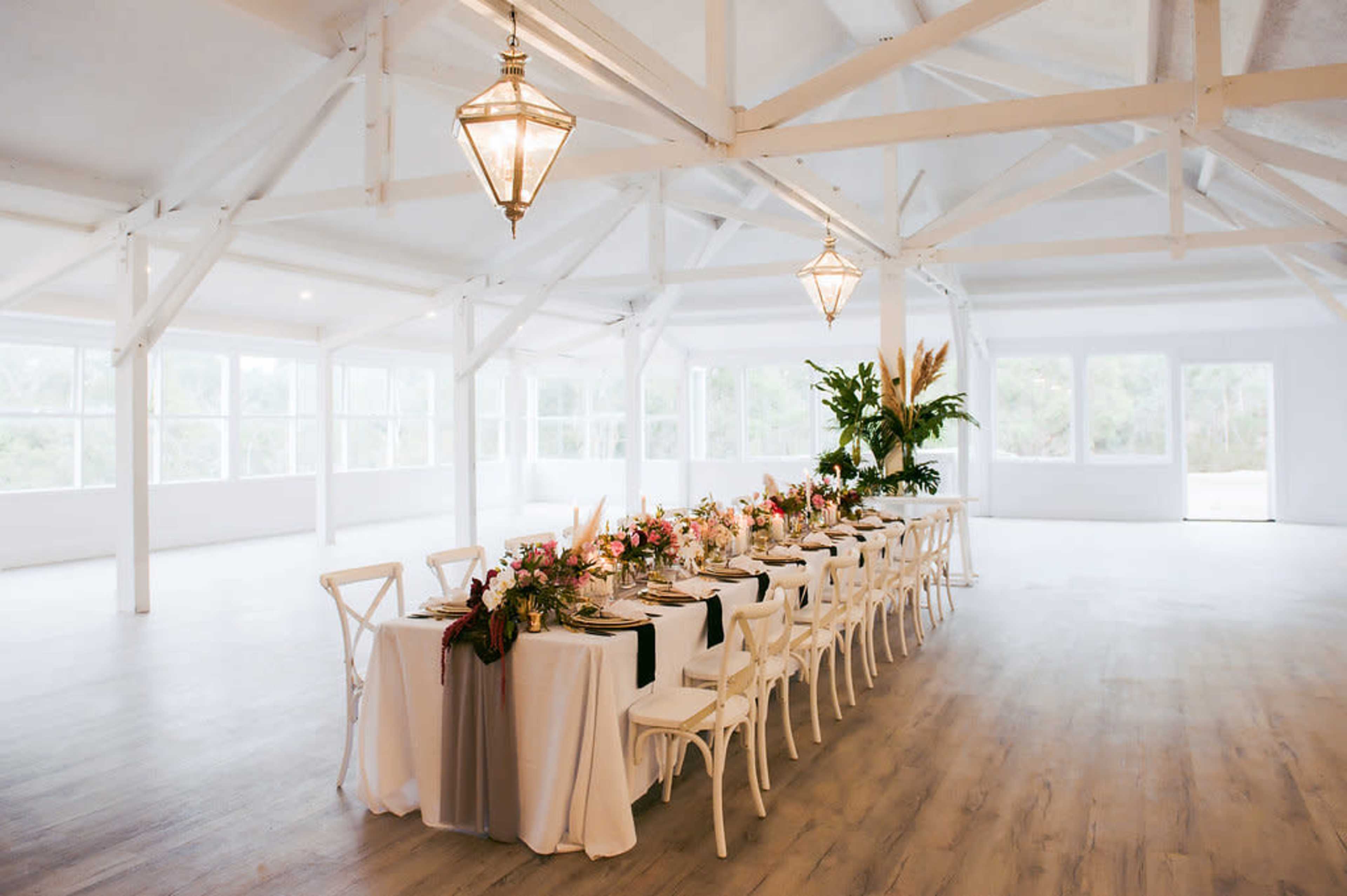 A long banquet table is elegantly set with floral centerpieces in a bright, airy room featuring large windows and exposed beams.