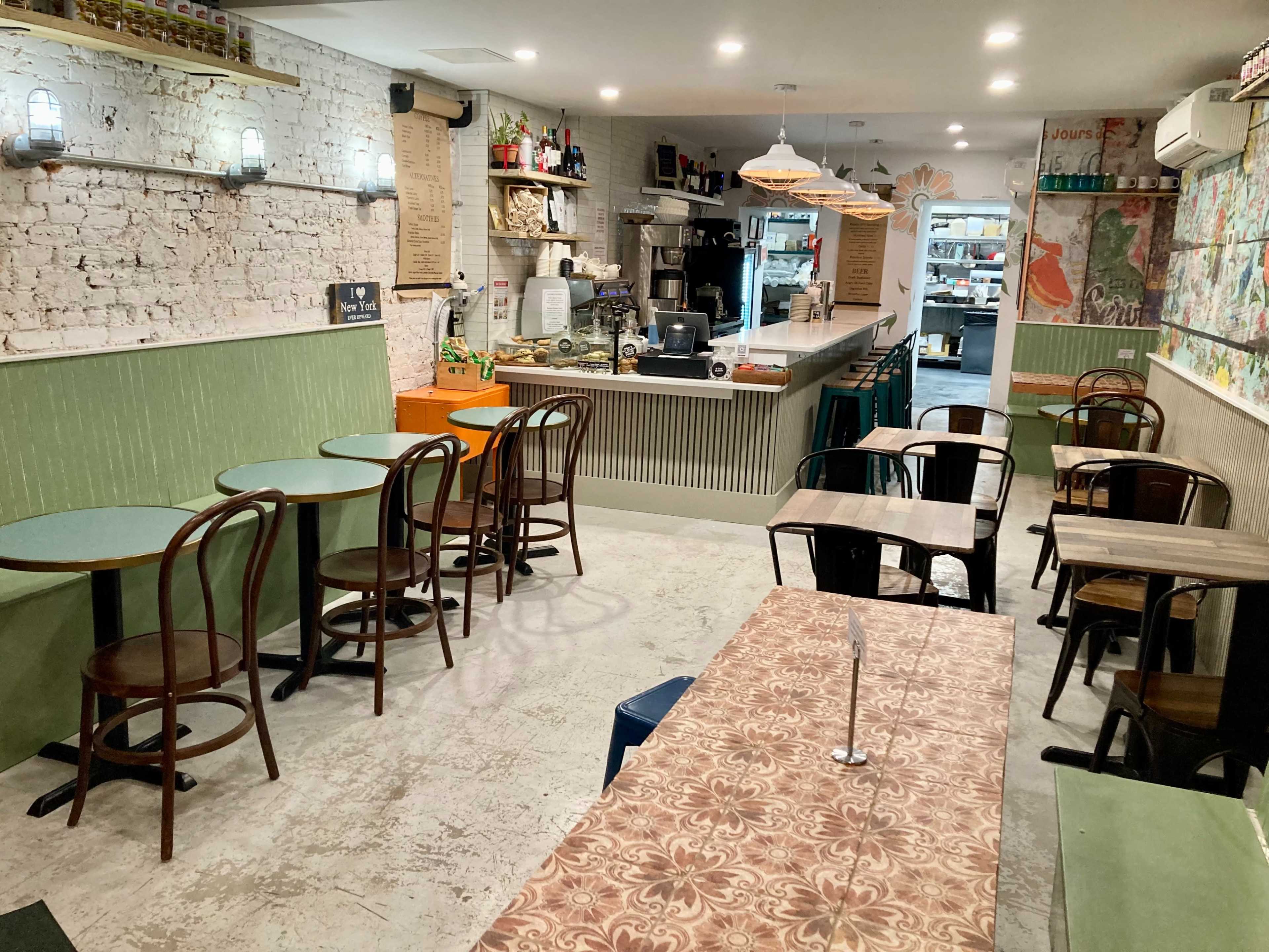 The image shows a small, modern cafe with wooden tables and chairs, a service counter in the background, and a menu displayed on the wall.
