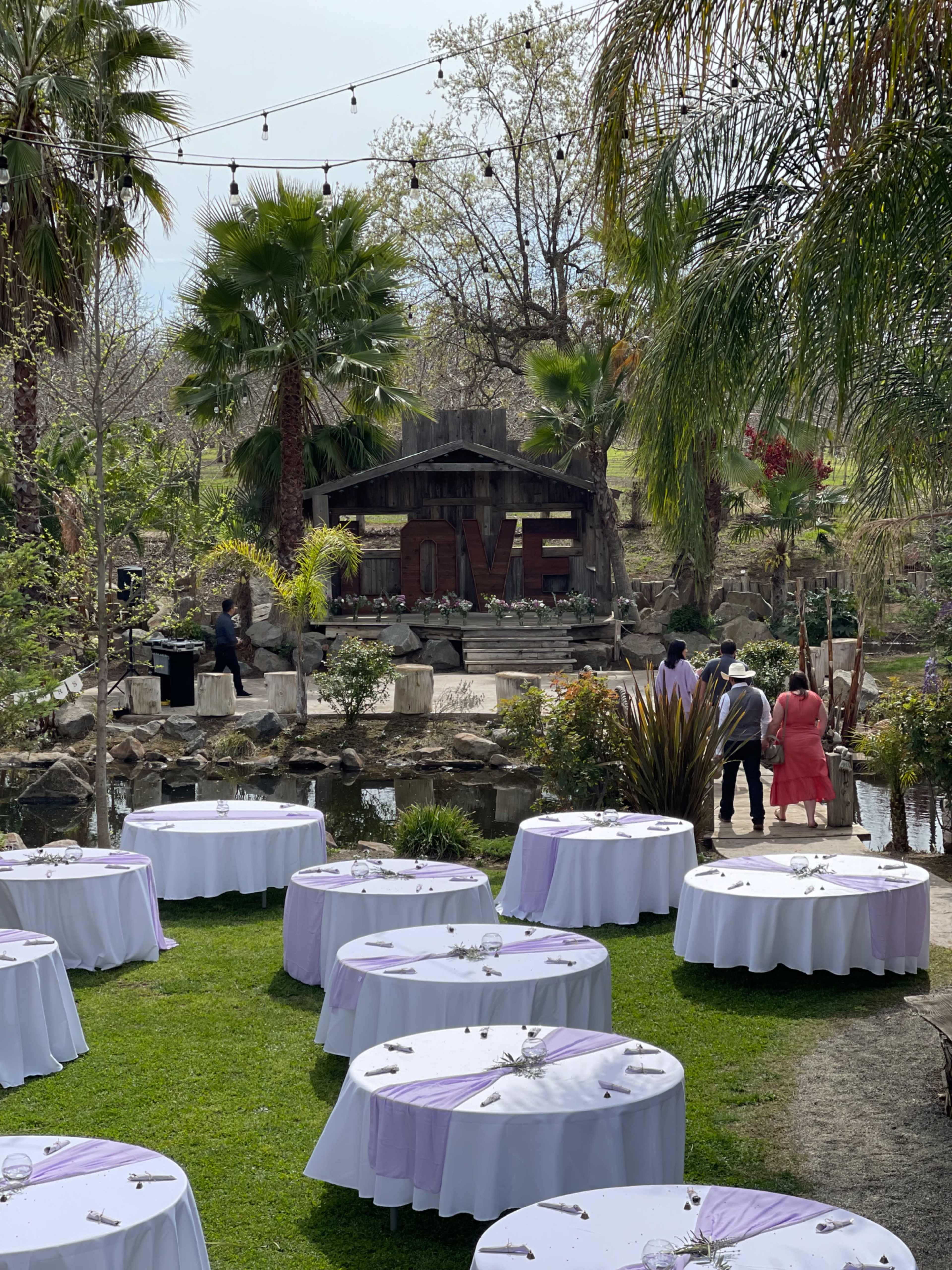 A garden venue is set up for an outdoor event, featuring round tables with lavender tablecloths and an ornate backdrop with a wooden structure surrounded by palm trees and a body of water.