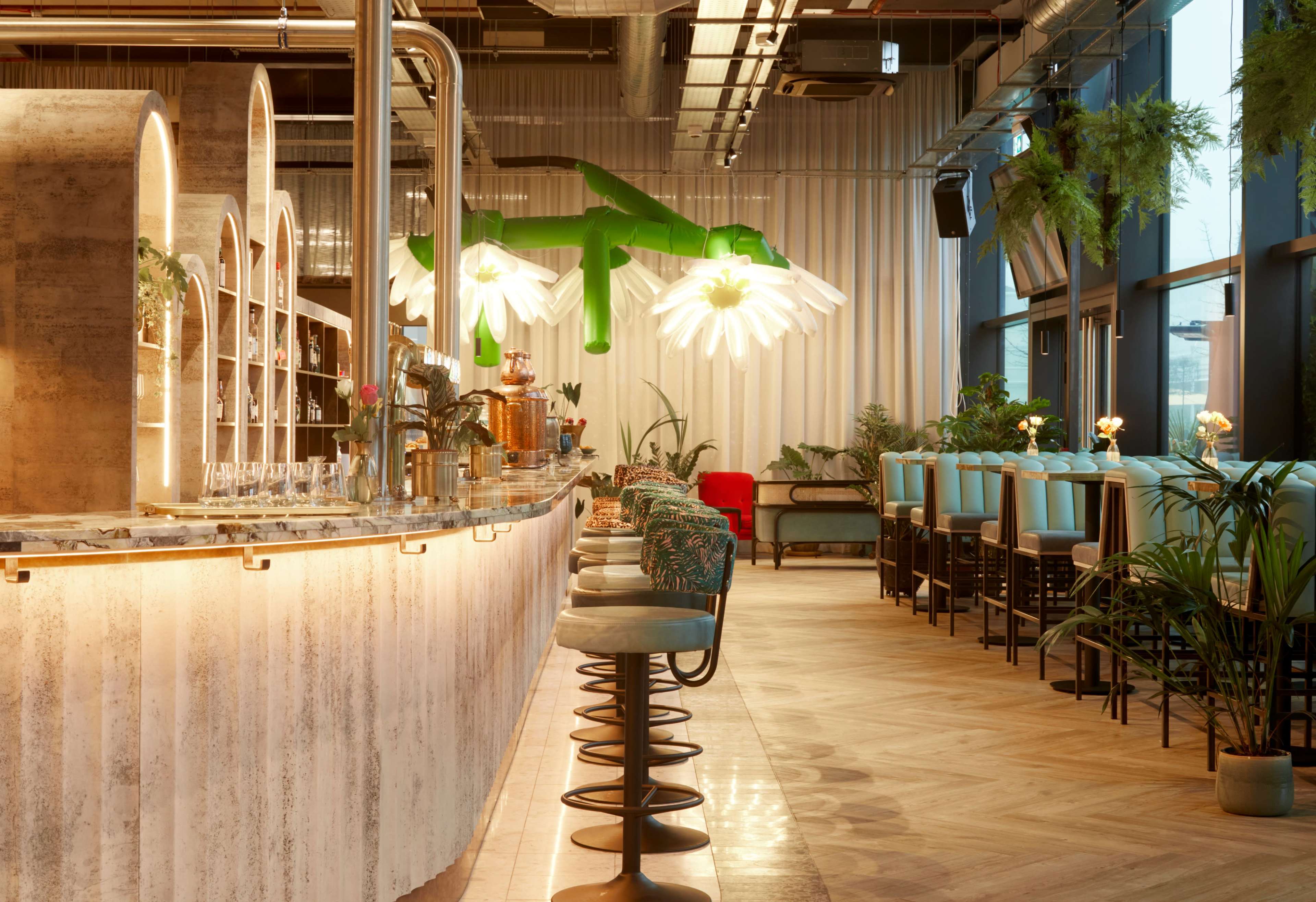 The image shows a modern bar area with a long marble countertop, high stools, and large pendant lights surrounded by indoor plants and seating arrangements.