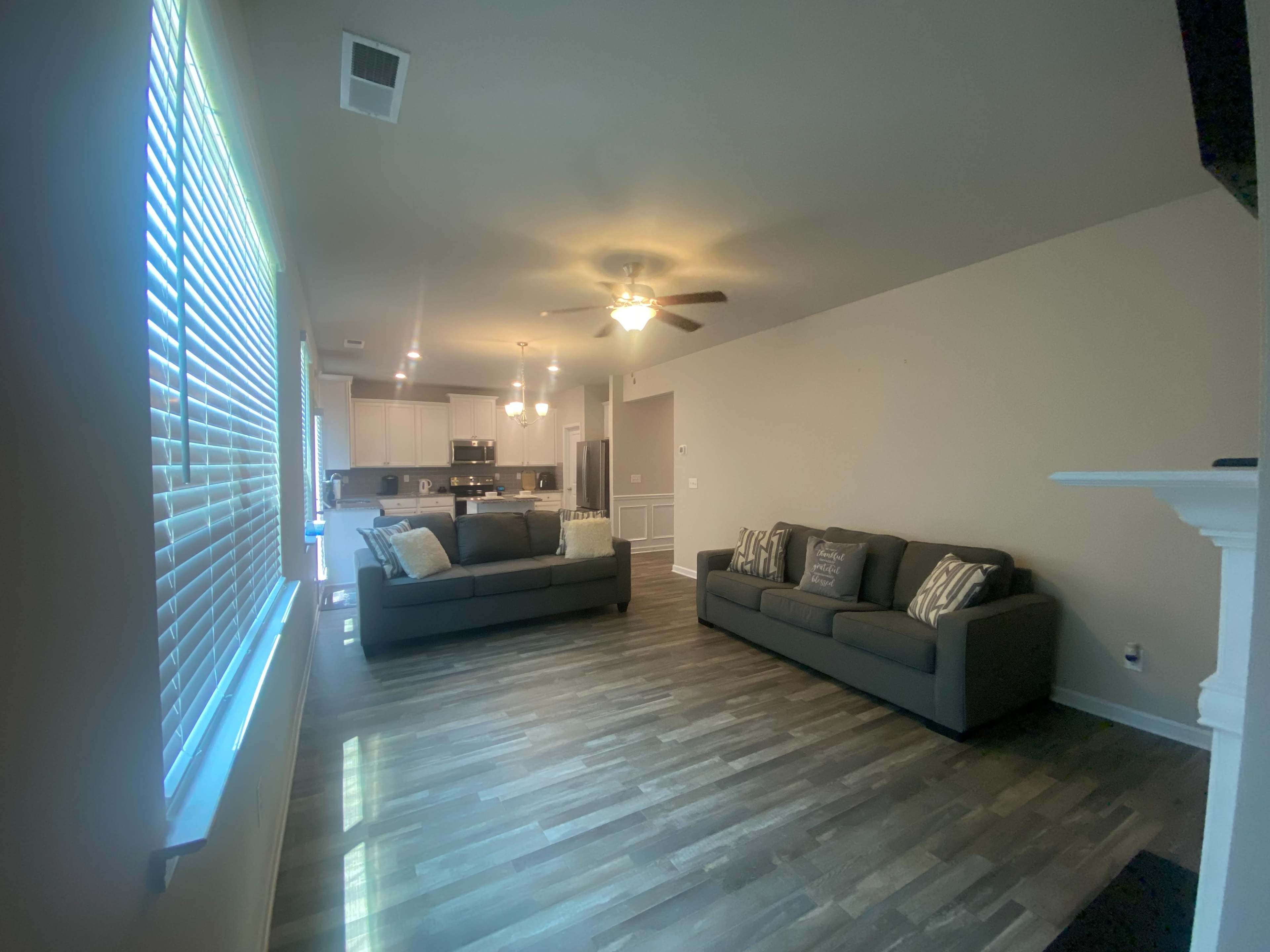 A living room with two gray sofas, large windows with blinds, and an open kitchen area in the background.