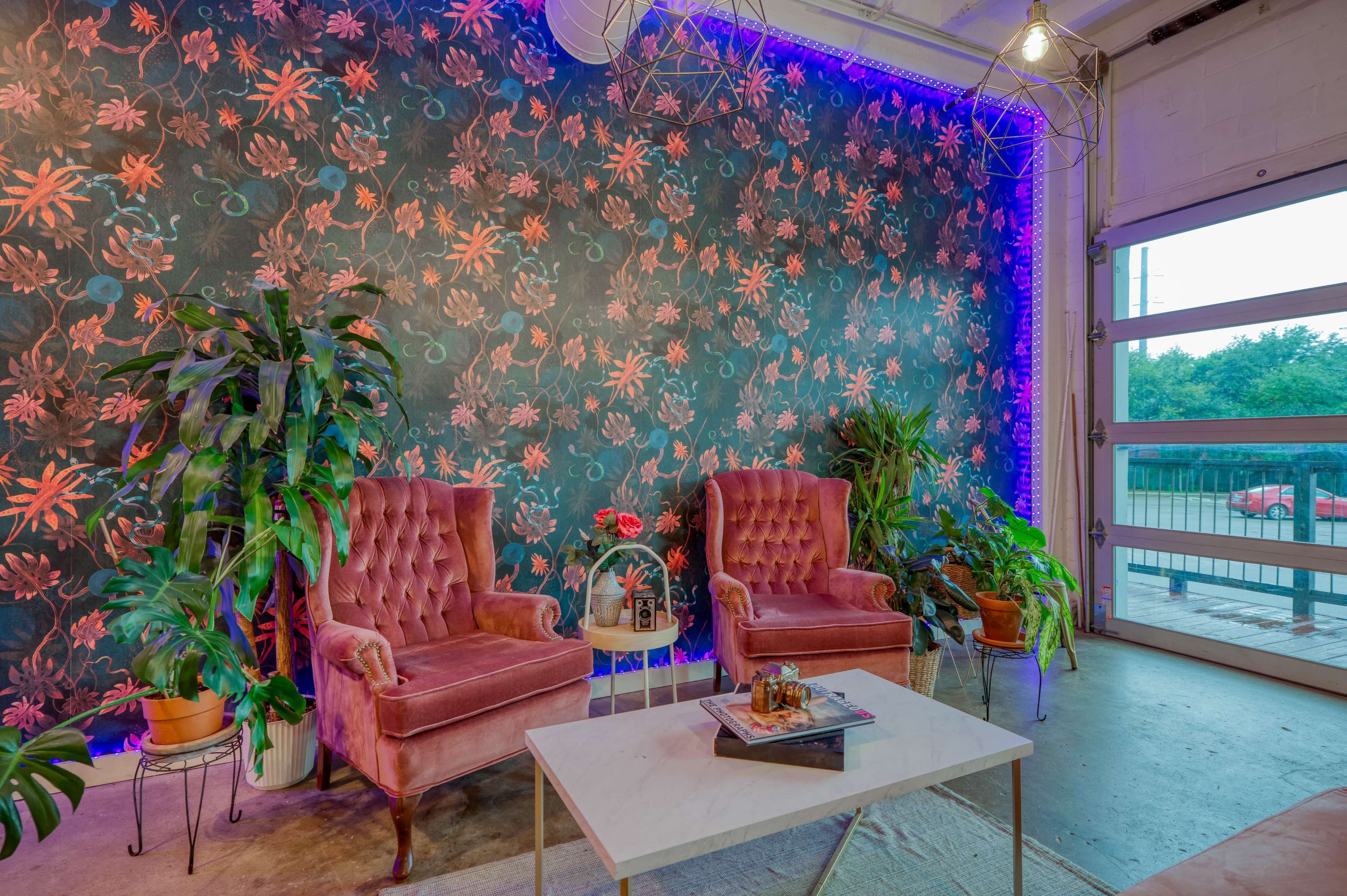 Two velvet armchairs are positioned beside a coffee table in front of a floral wallpaper backdrop, with indoor plants placed around the scene.