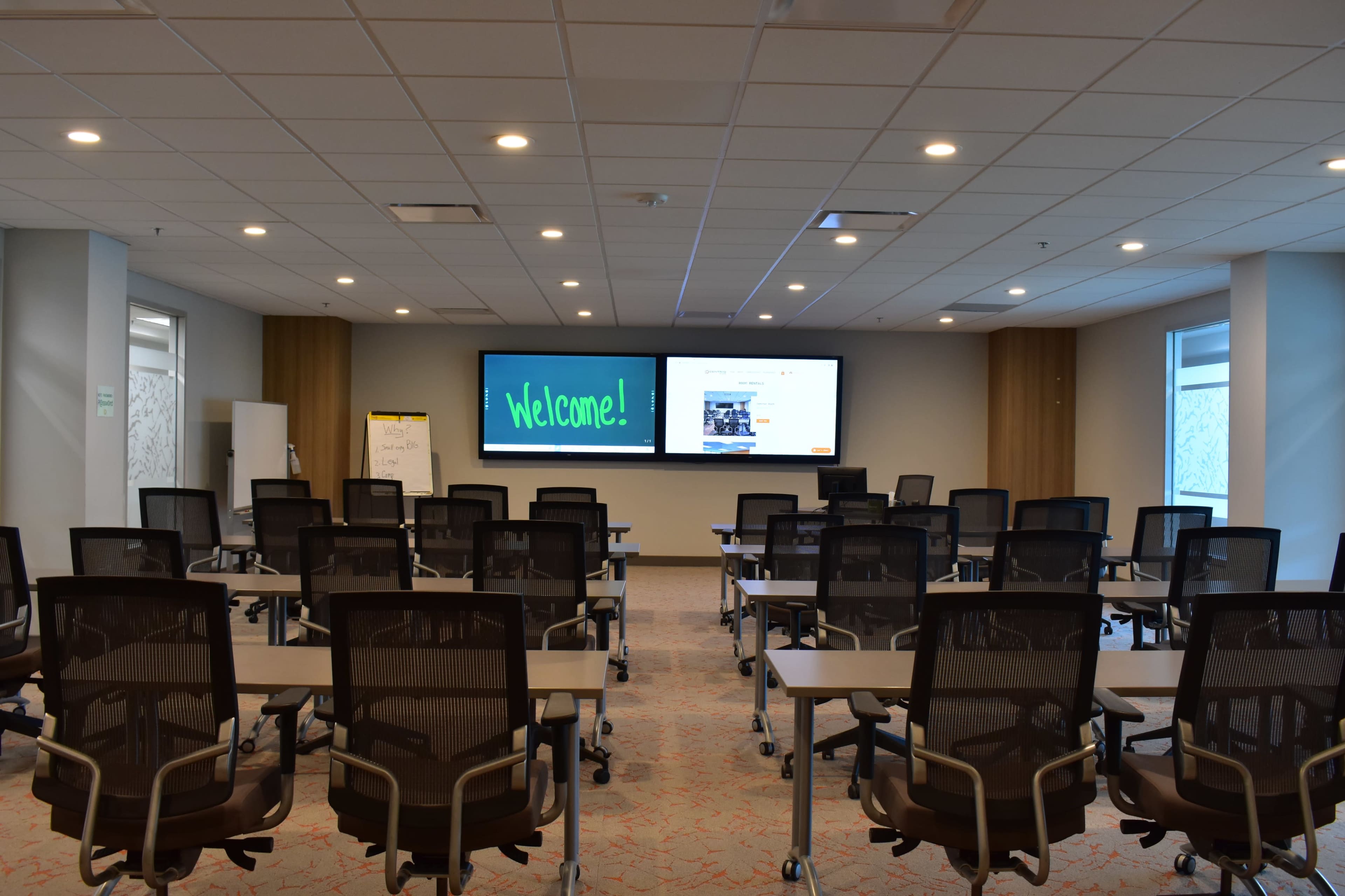 A modern conference room is set up with rows of desks and chairs facing a large screen displaying a welcome message.