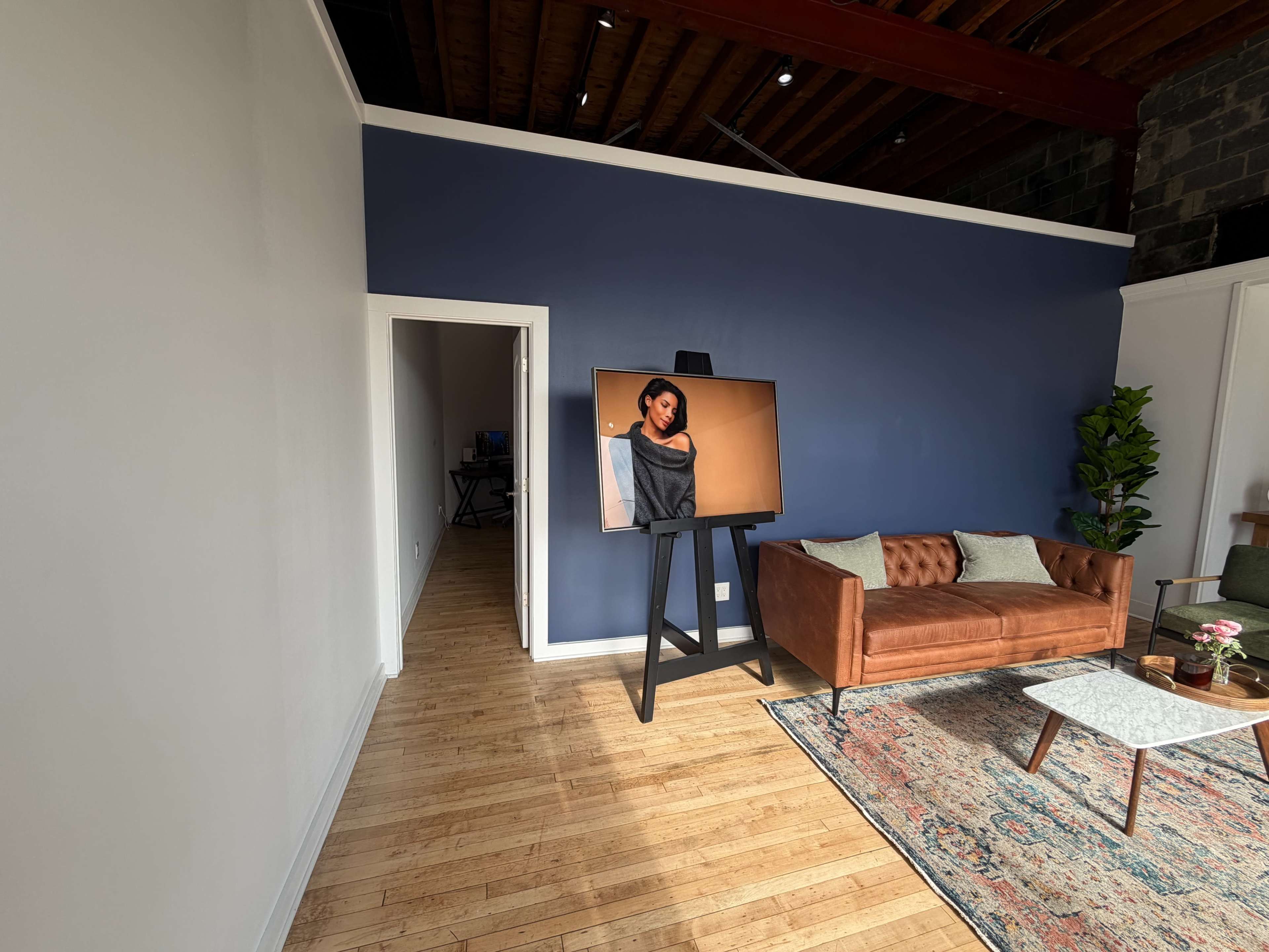 The image shows a spacious room featuring a brown leather sofa, a coffee table, and a large artwork displayed on an easel against a deep blue wall.