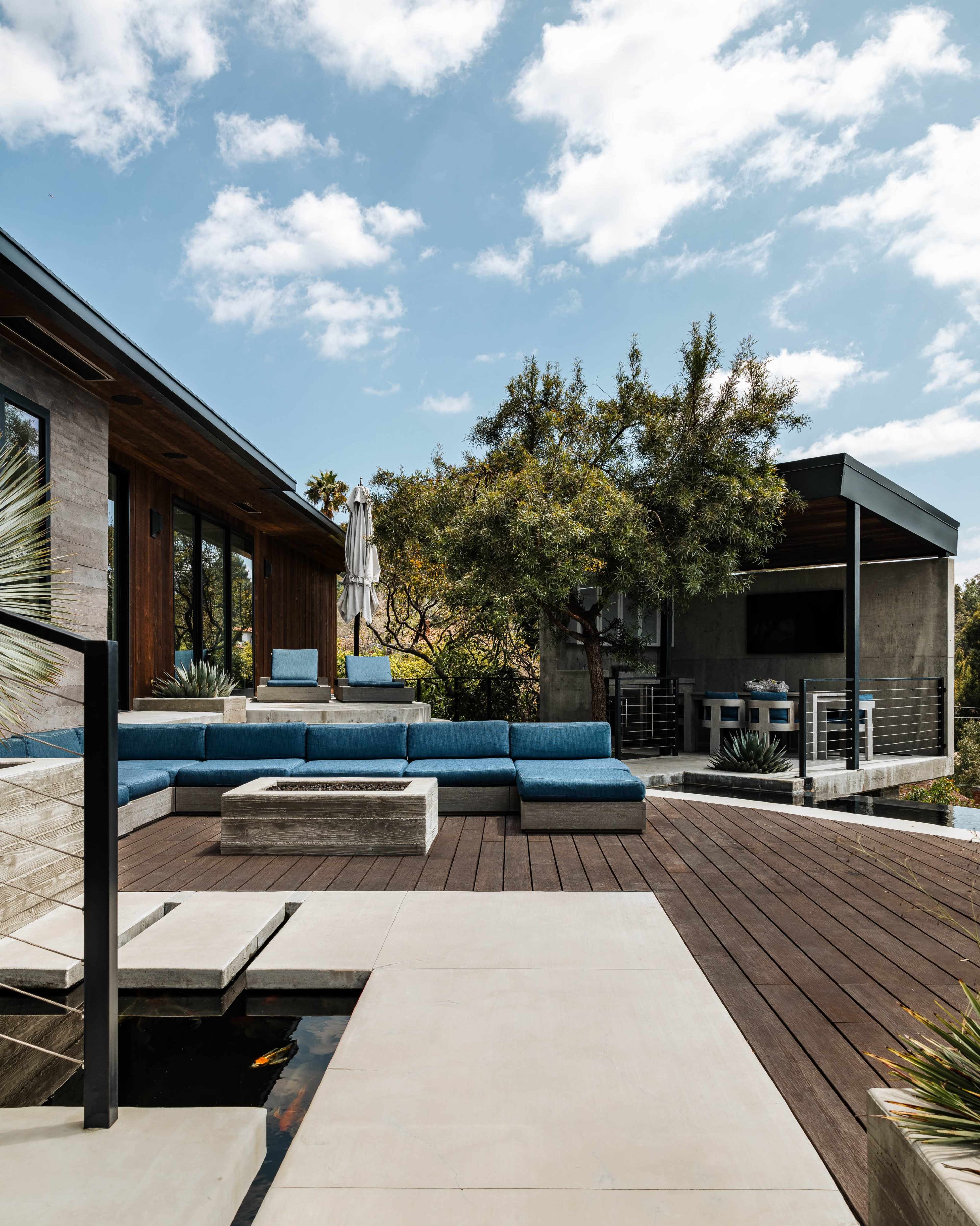The image shows a modern outdoor patio area with blue seating, a fire pit, and a nearby shaded structure in front of a landscaped yard.