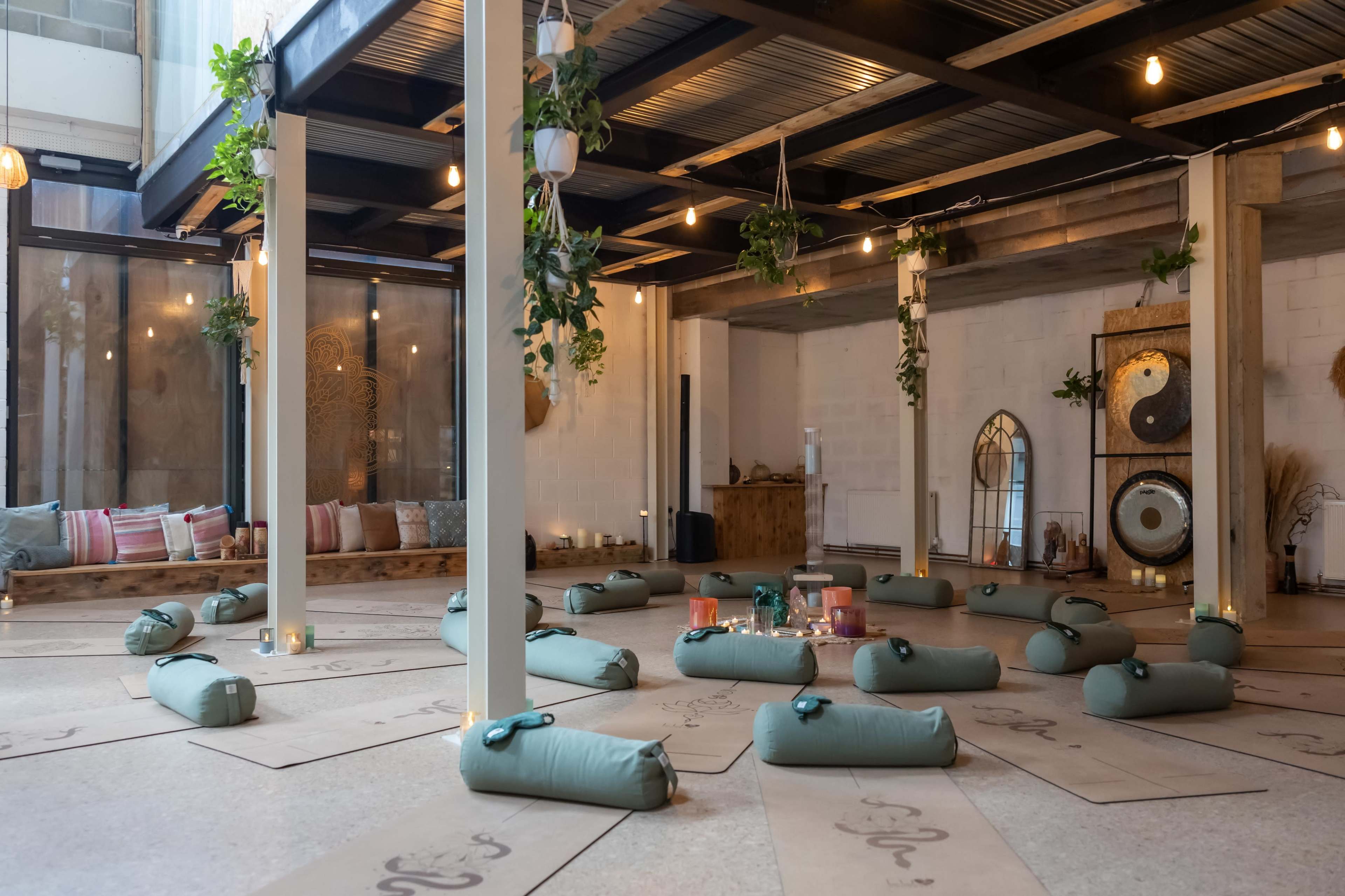 The image shows a spacious indoor studio set up for a yoga or meditation session, featuring yoga mats placed on the floor, decorative plants hanging from the ceiling, and ambient lighting.