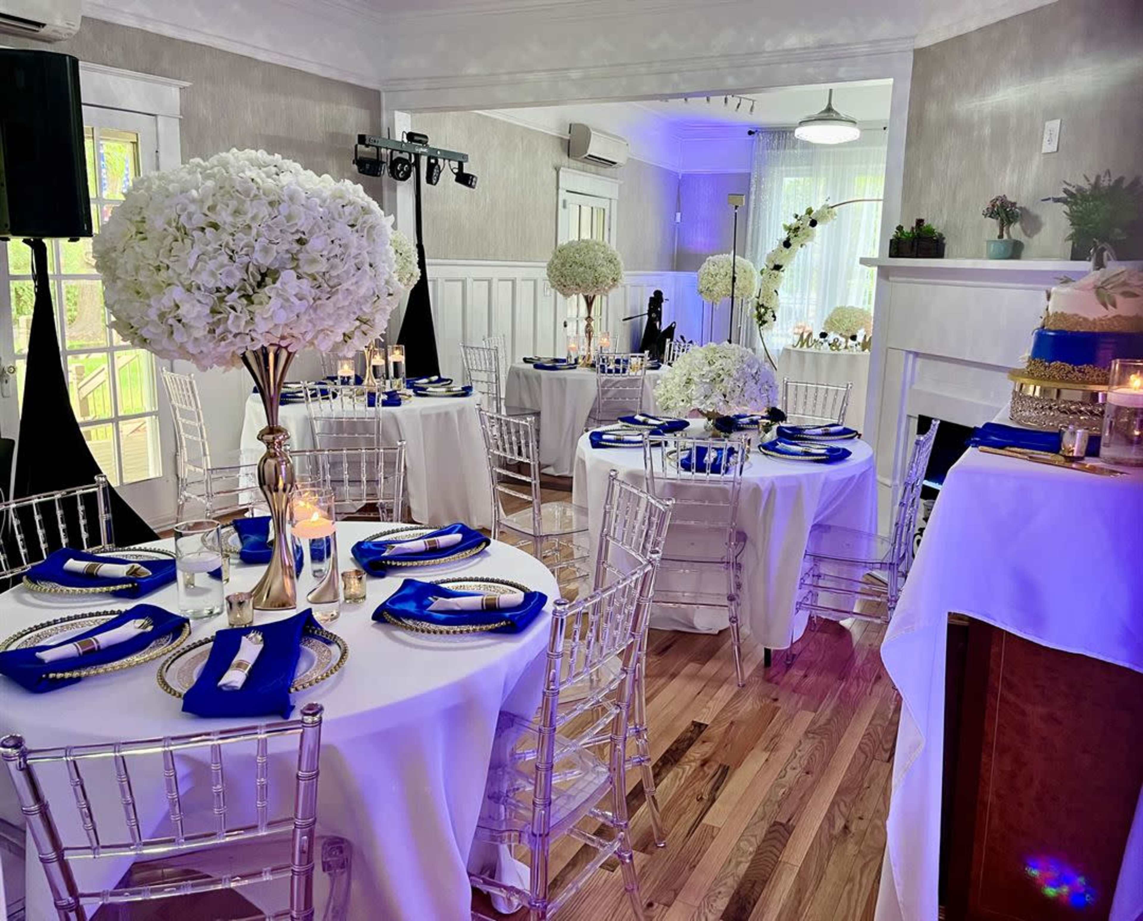 A decorated event space with round tables covered in white tablecloths, each featuring a tall centerpiece of white flowers and blue napkins, set up for a formal gathering.