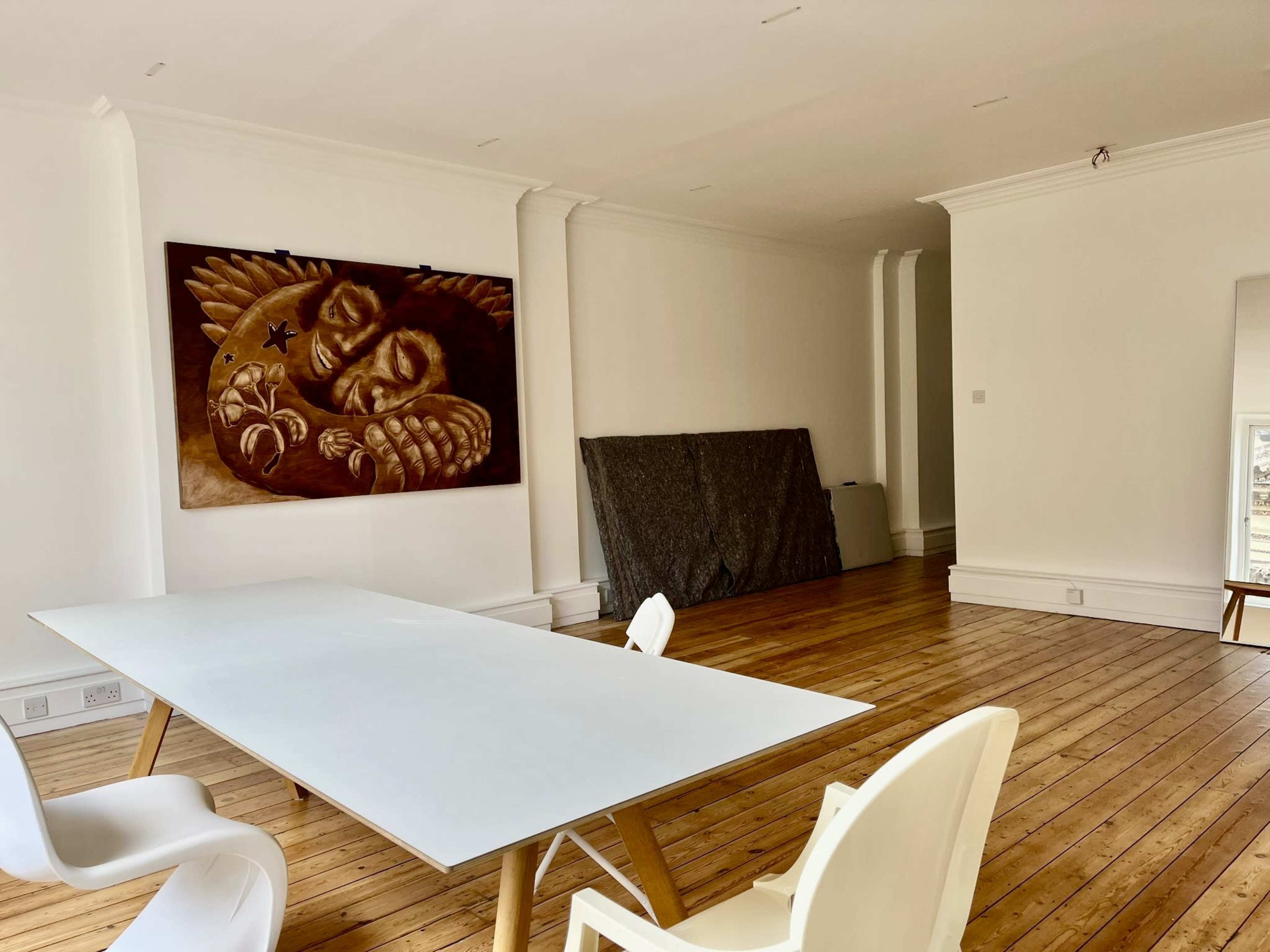 A spacious room with wooden floors, featuring a large table, two white chairs, and a prominent mural of a face with wings on the wall.