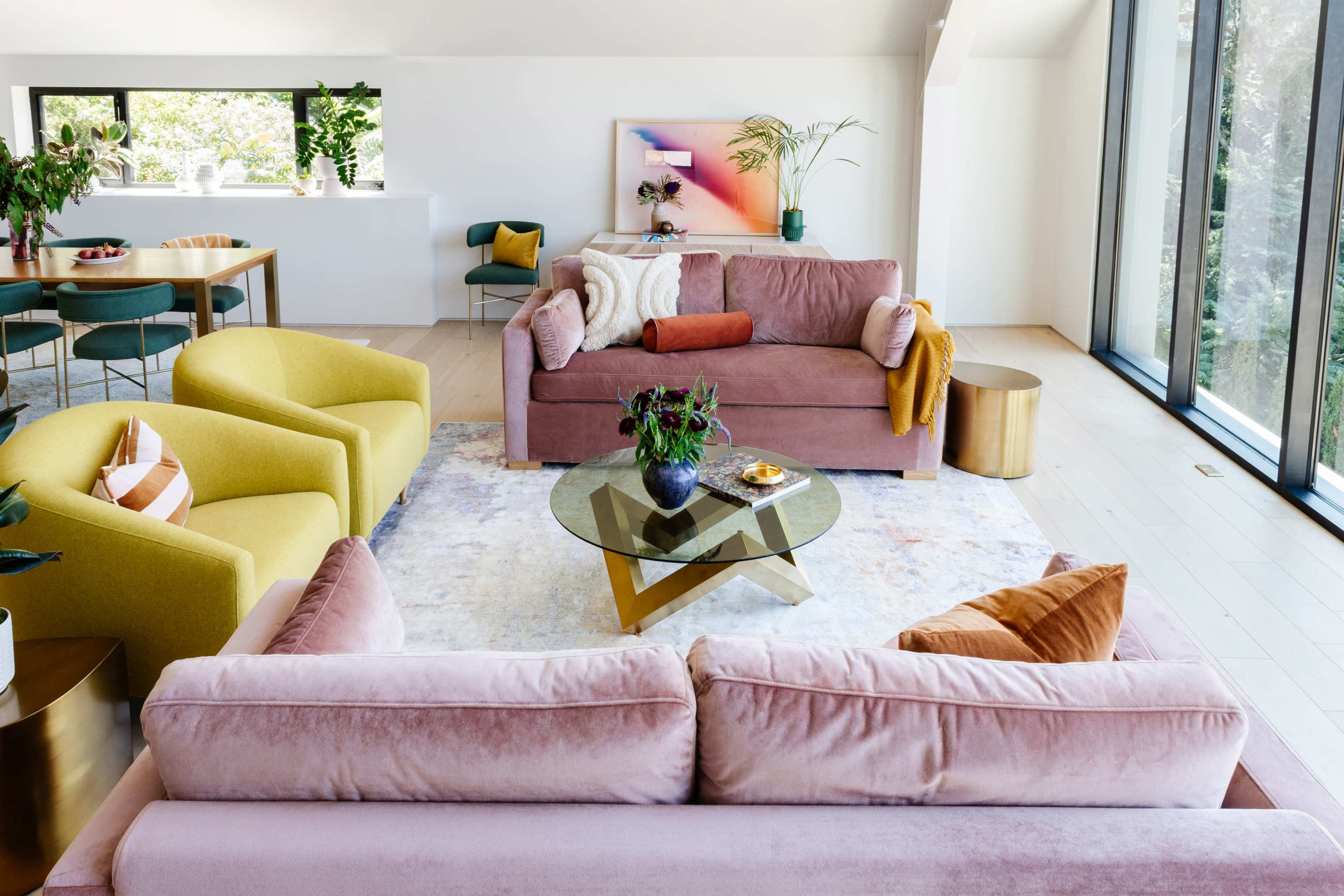 A modern living room features two couches, colorful armchairs, a glass coffee table, and large windows letting in natural light.