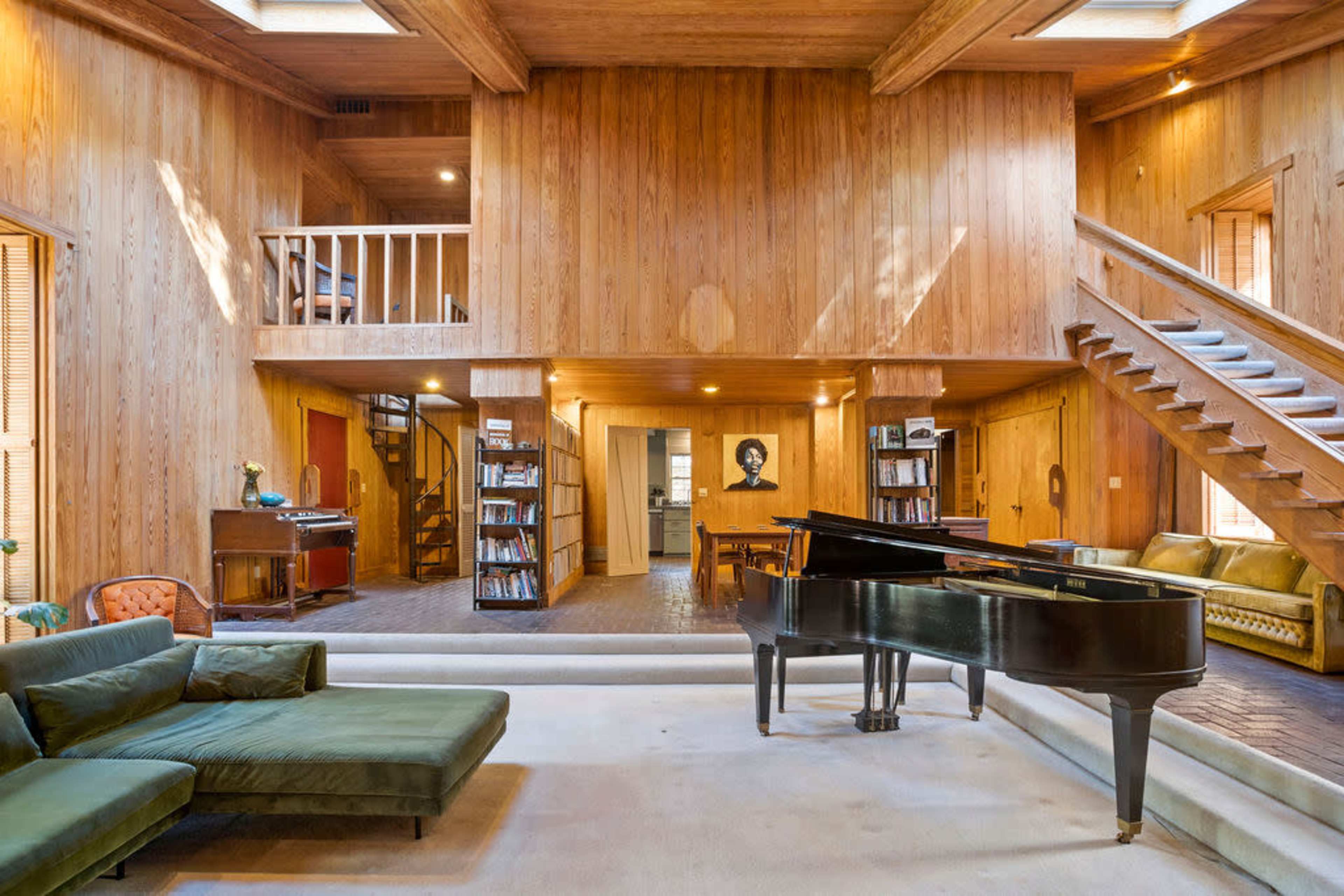 A spacious, wooden-paneled living area featuring a grand piano, a staircase, and a second-floor balcony overlooking the main space.