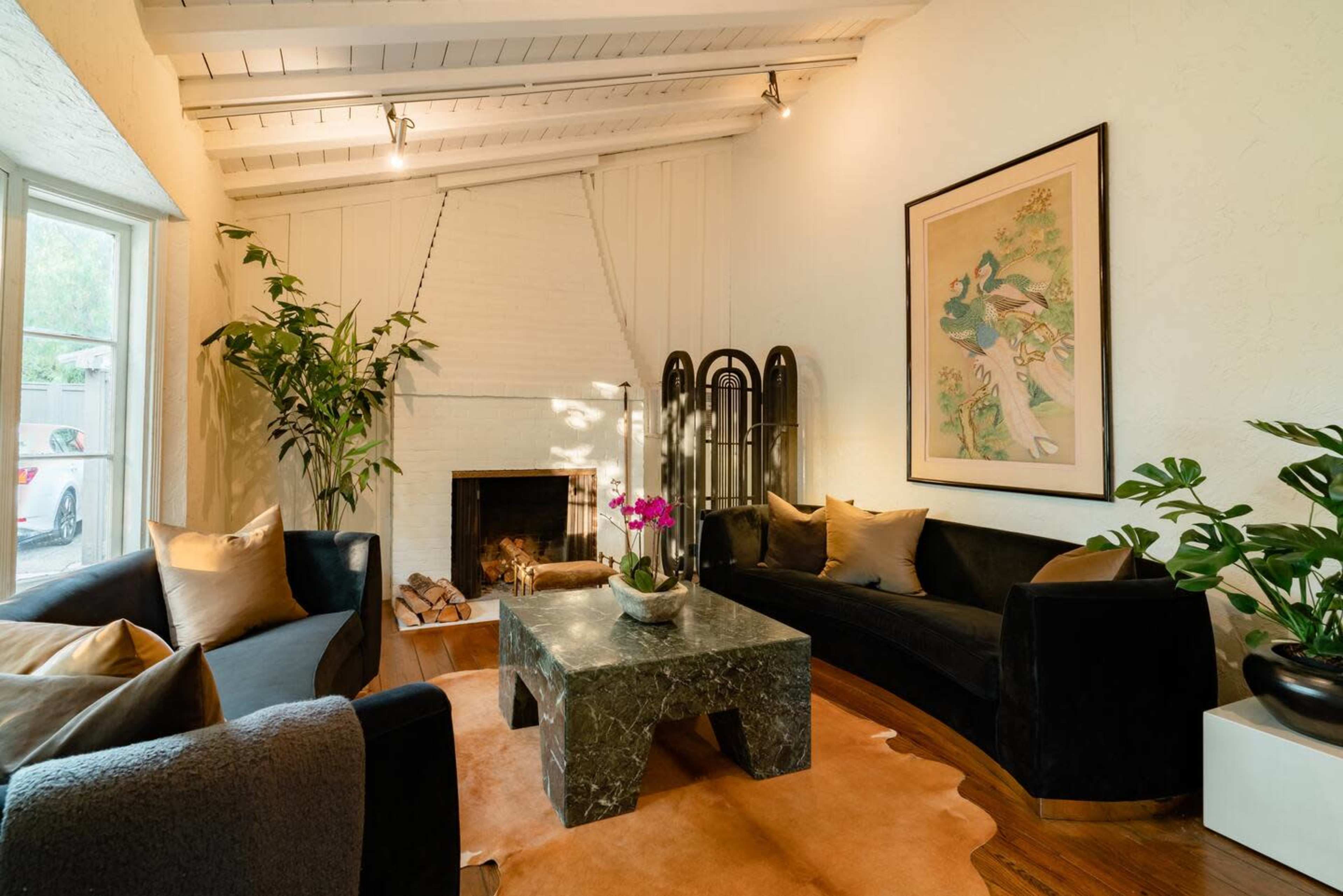 The image shows a stylish living room featuring a fireplace, a large green marble coffee table, and a mix of seating arrangements with plants and artwork on the walls.