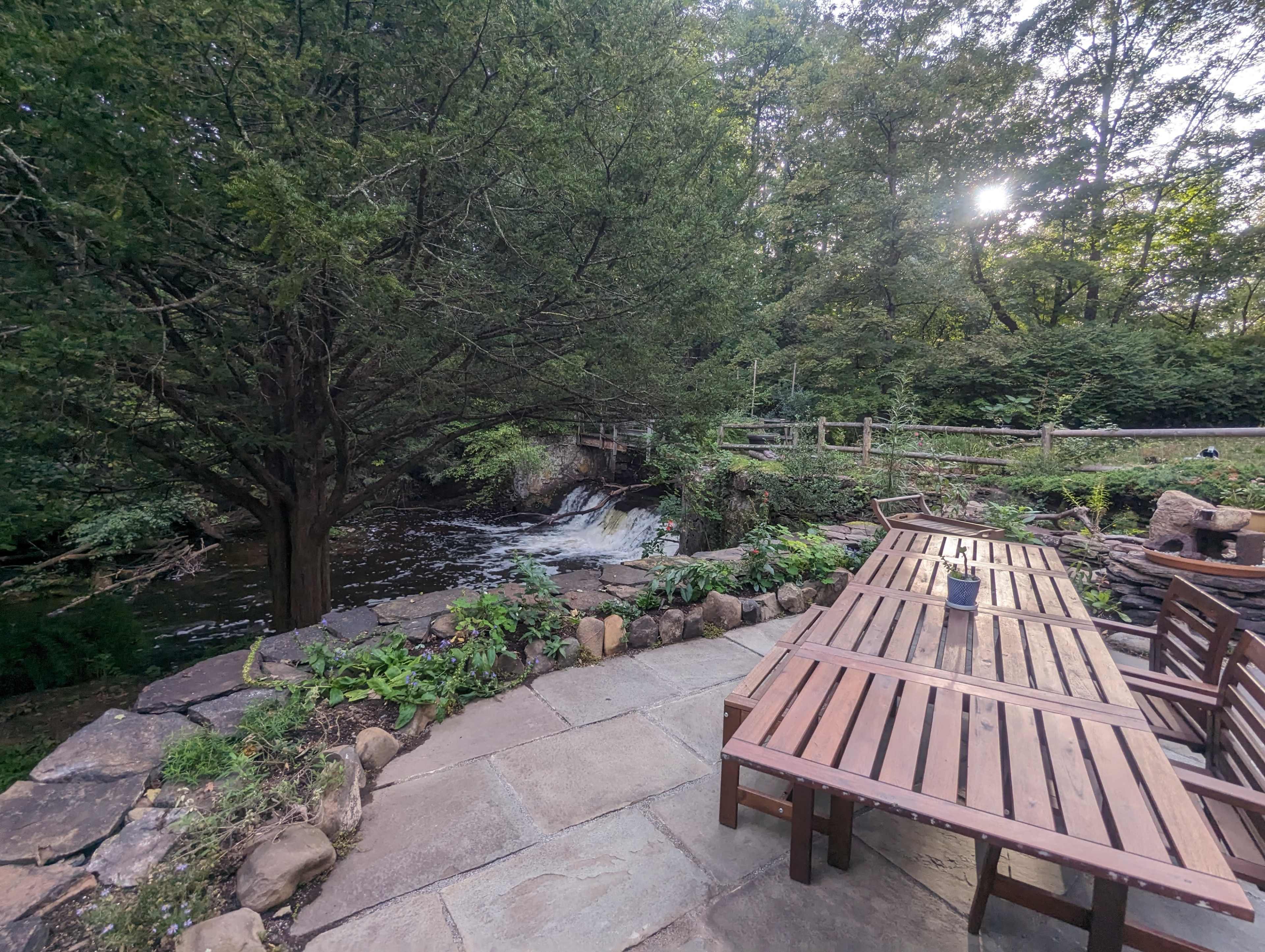A wooden table and benches are set along a stone path near a flowing stream surrounded by trees.