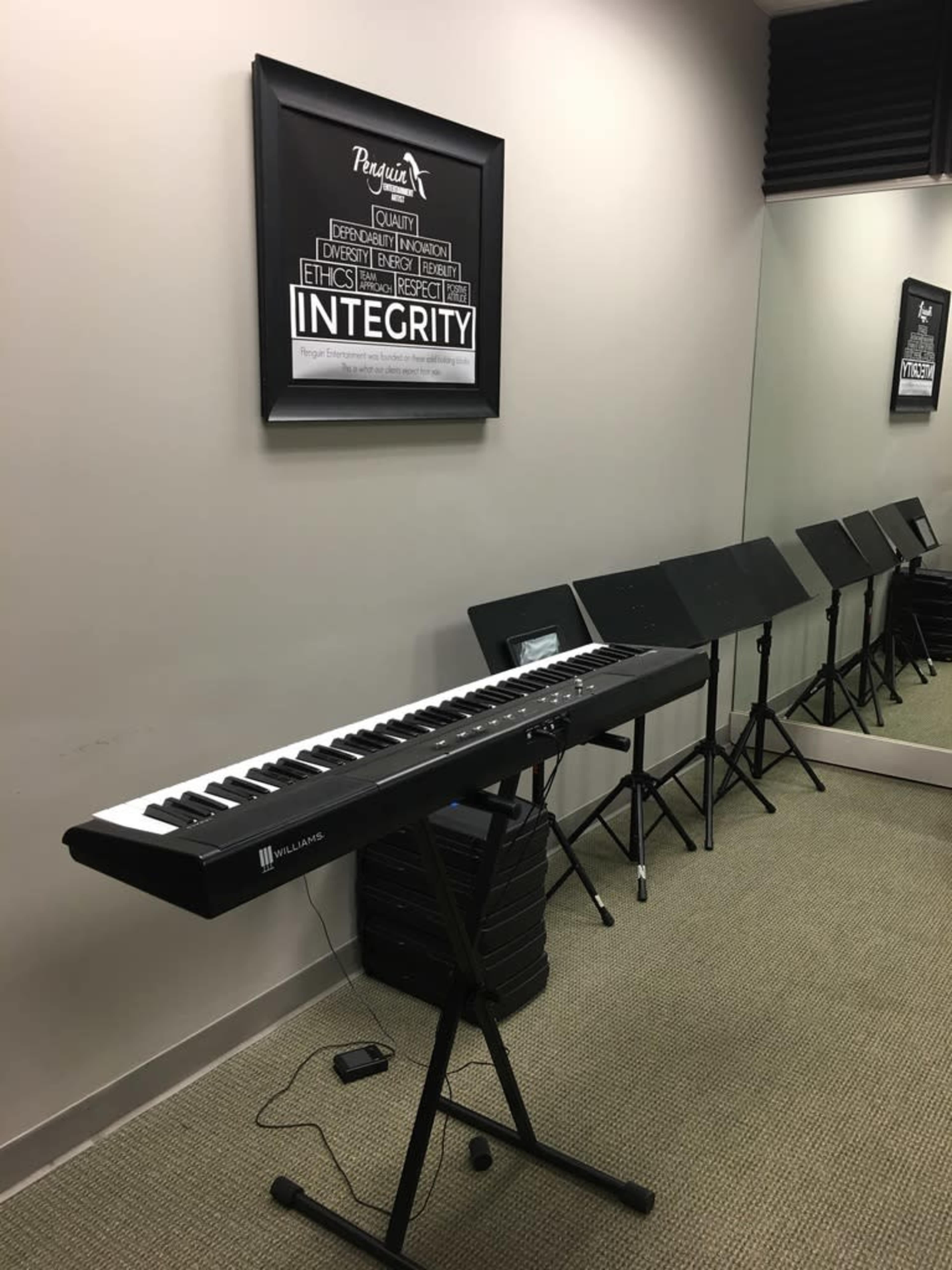 A digital keyboard on a stand in a music practice room, with sheet music stands lined up against the wall and a framed poster displaying the word "INTEGRITY."
