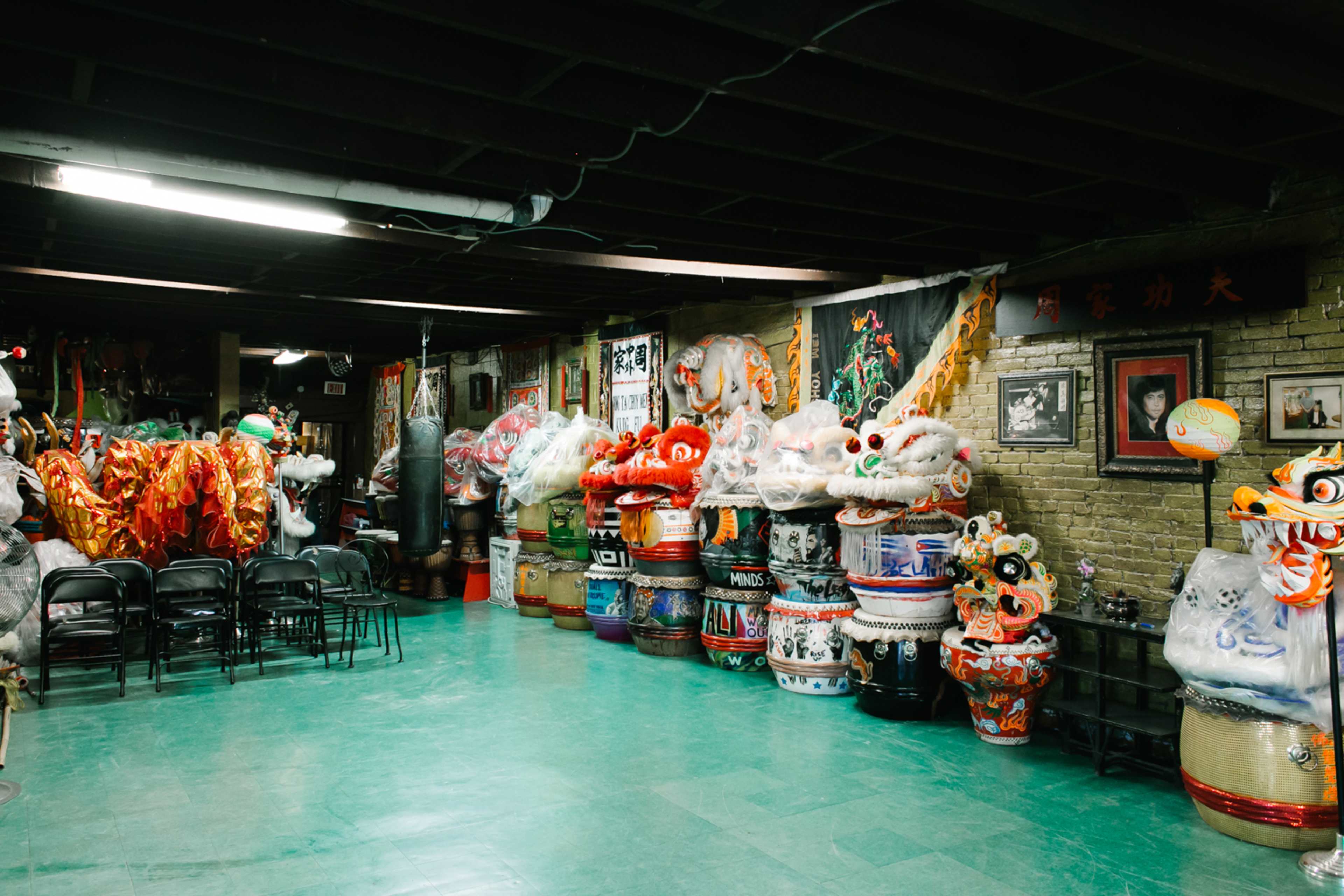 A dimly lit room filled with colorful dragon and lion dance costumes and masks along the walls and stacks of decorative drums.