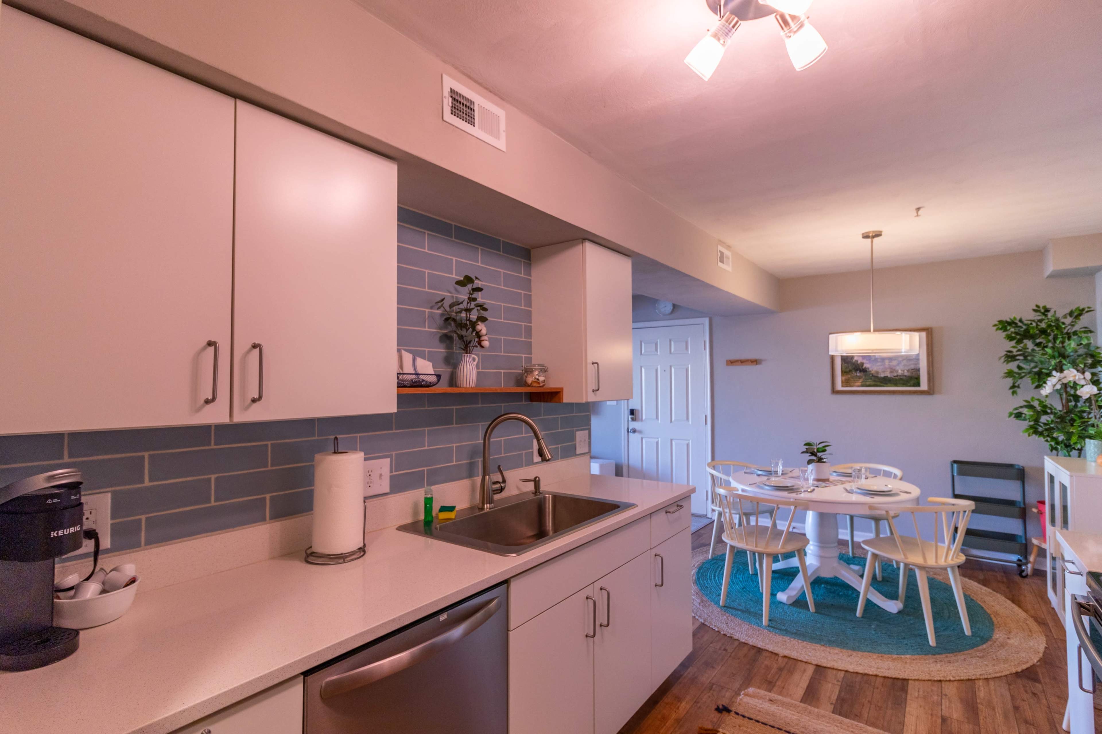 The image shows a modern kitchen with light-colored cabinets, a stainless steel sink, and a blue tiled backsplash, adjacent to a dining area with a round table and white chairs.