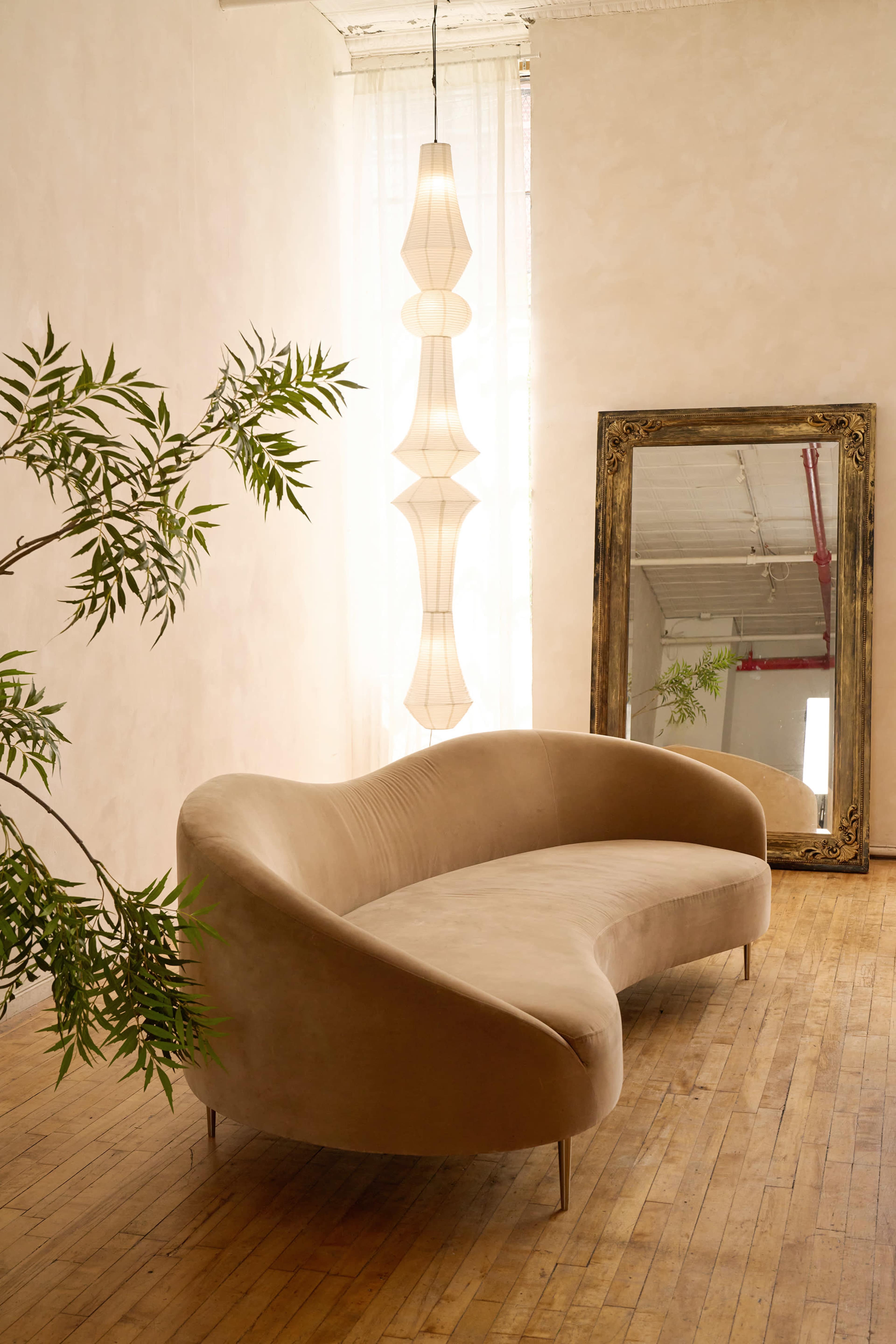 A curved beige sofa sits on a wooden floor beneath a large decorative mirror and a pendant light, with a plant nearby.