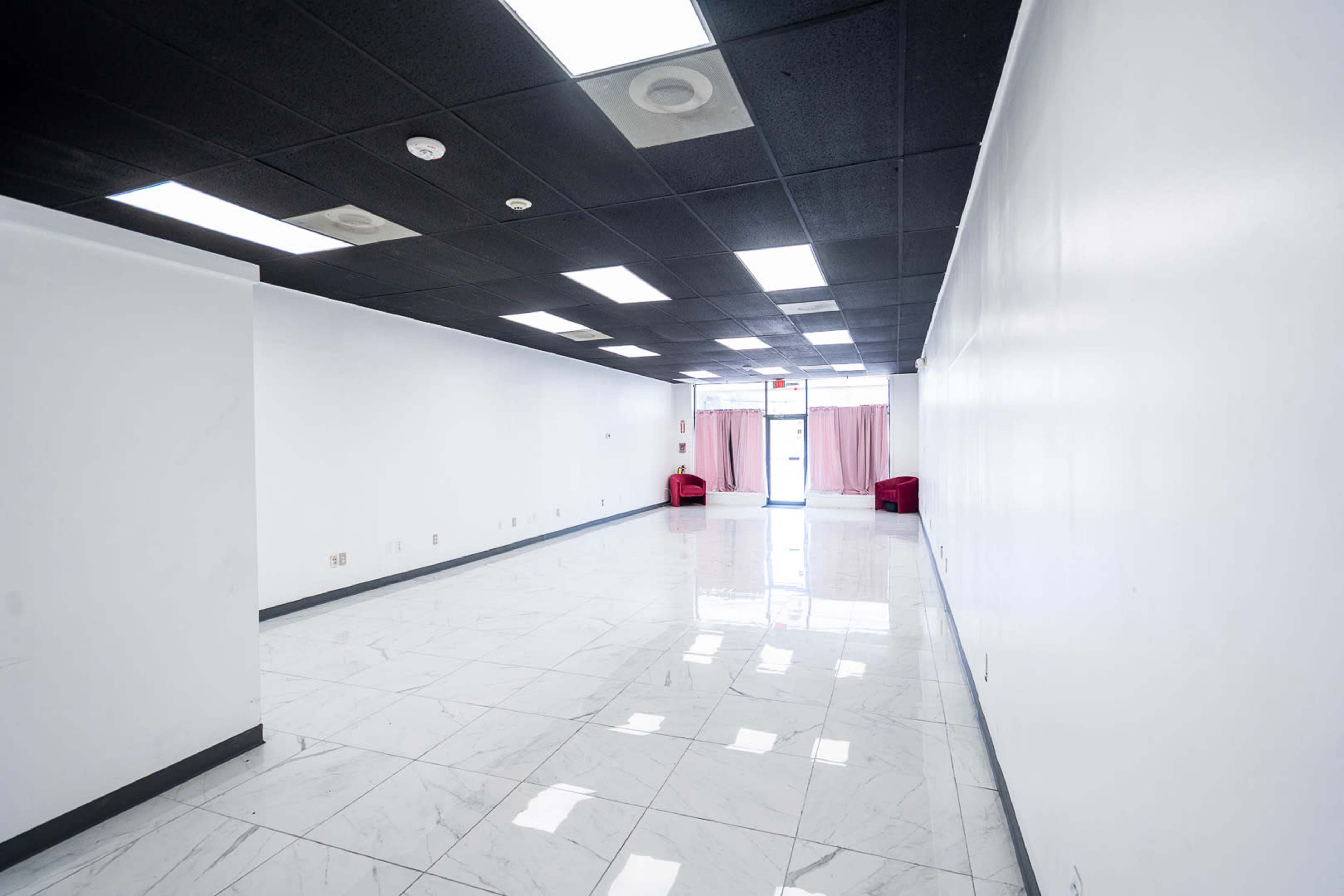 The image shows an empty interior space with white walls, a shiny tiled floor, and two red chairs near the entrance.