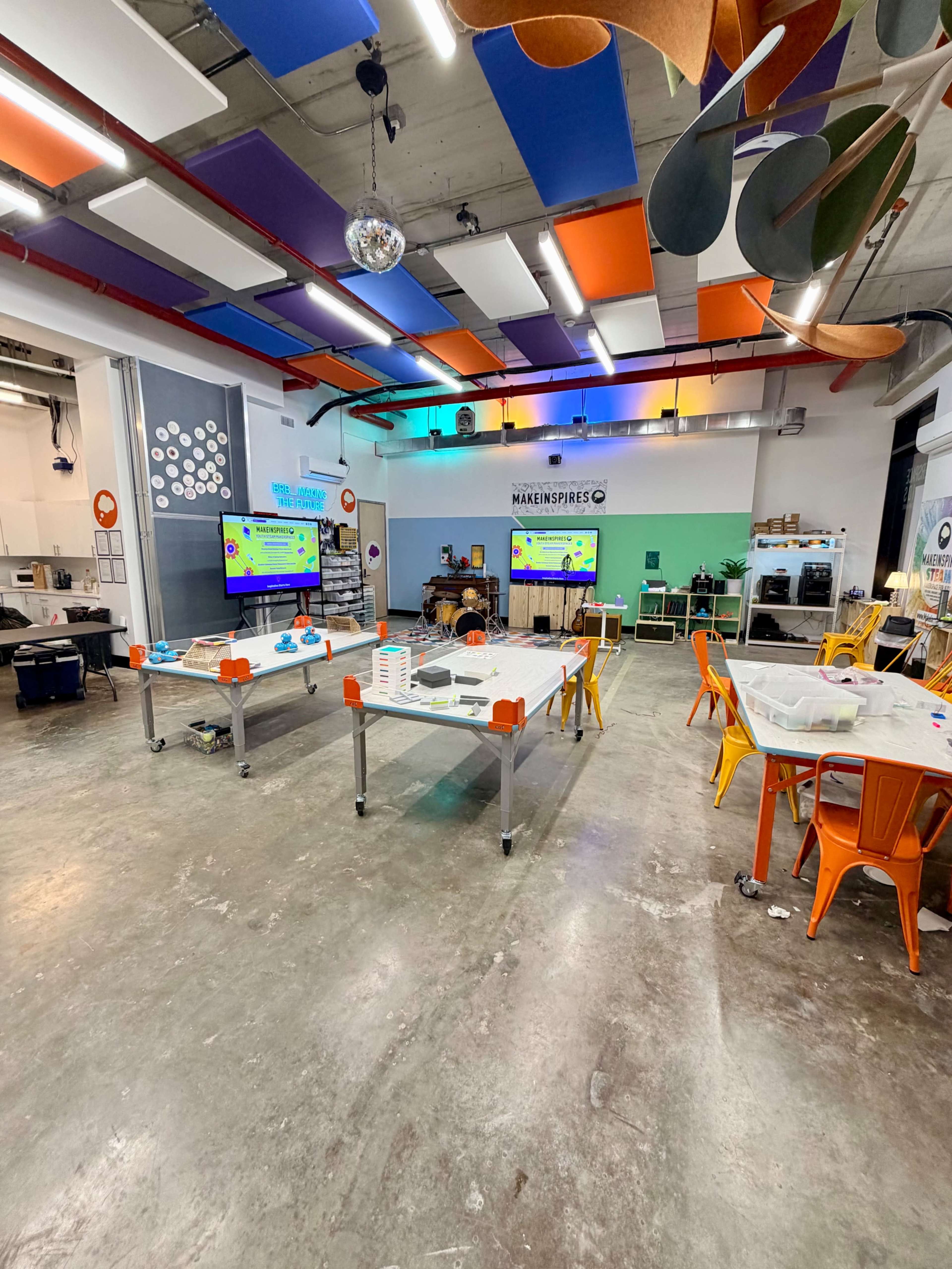 The image shows a modern classroom or workshop space with several tables, colorful chairs, and multiple screens displaying information on the walls, featuring diverse ceiling panels.