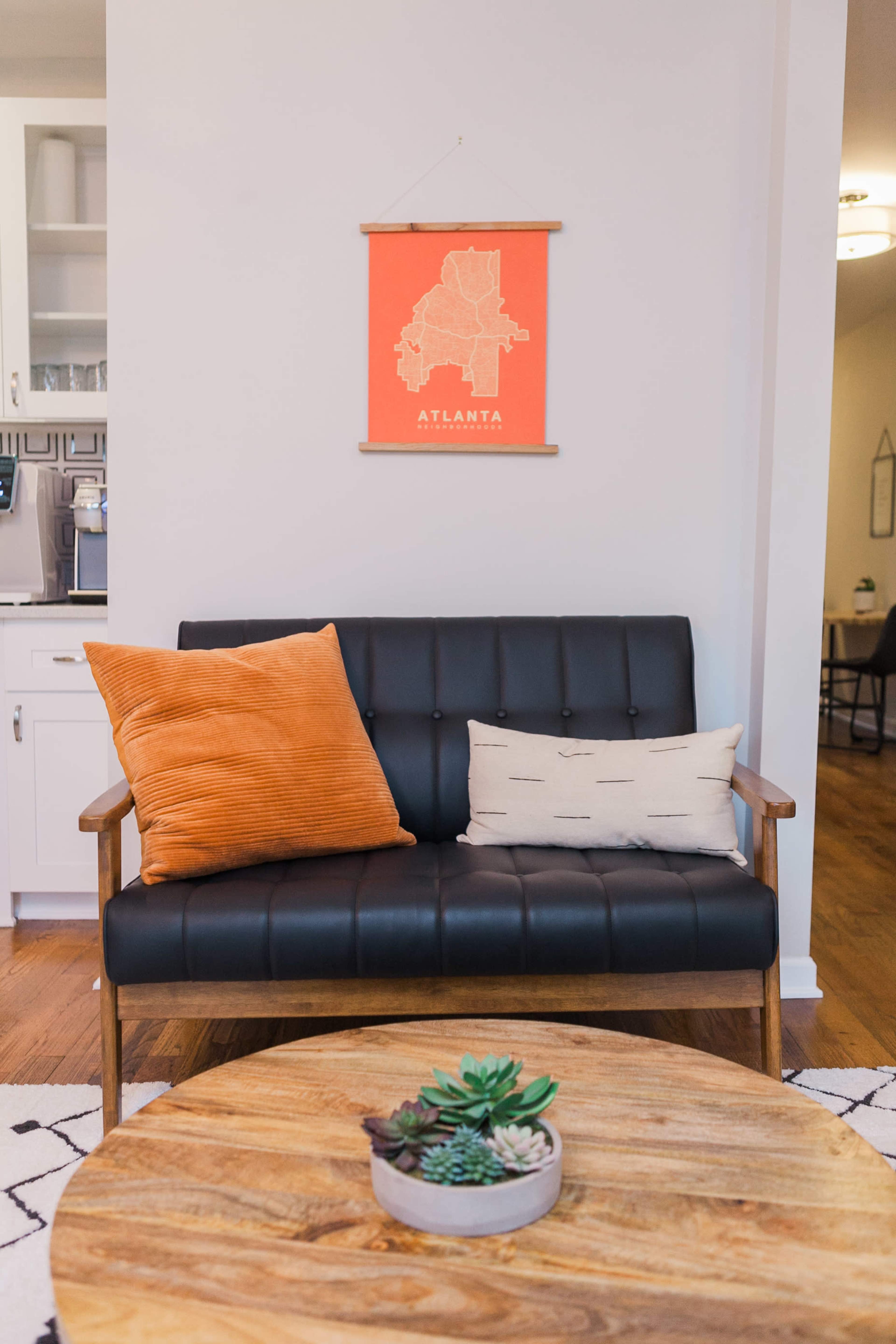 A black leather couch with orange and cream pillows is positioned in front of a round wooden coffee table, under a wall featuring an orange map of Atlanta.