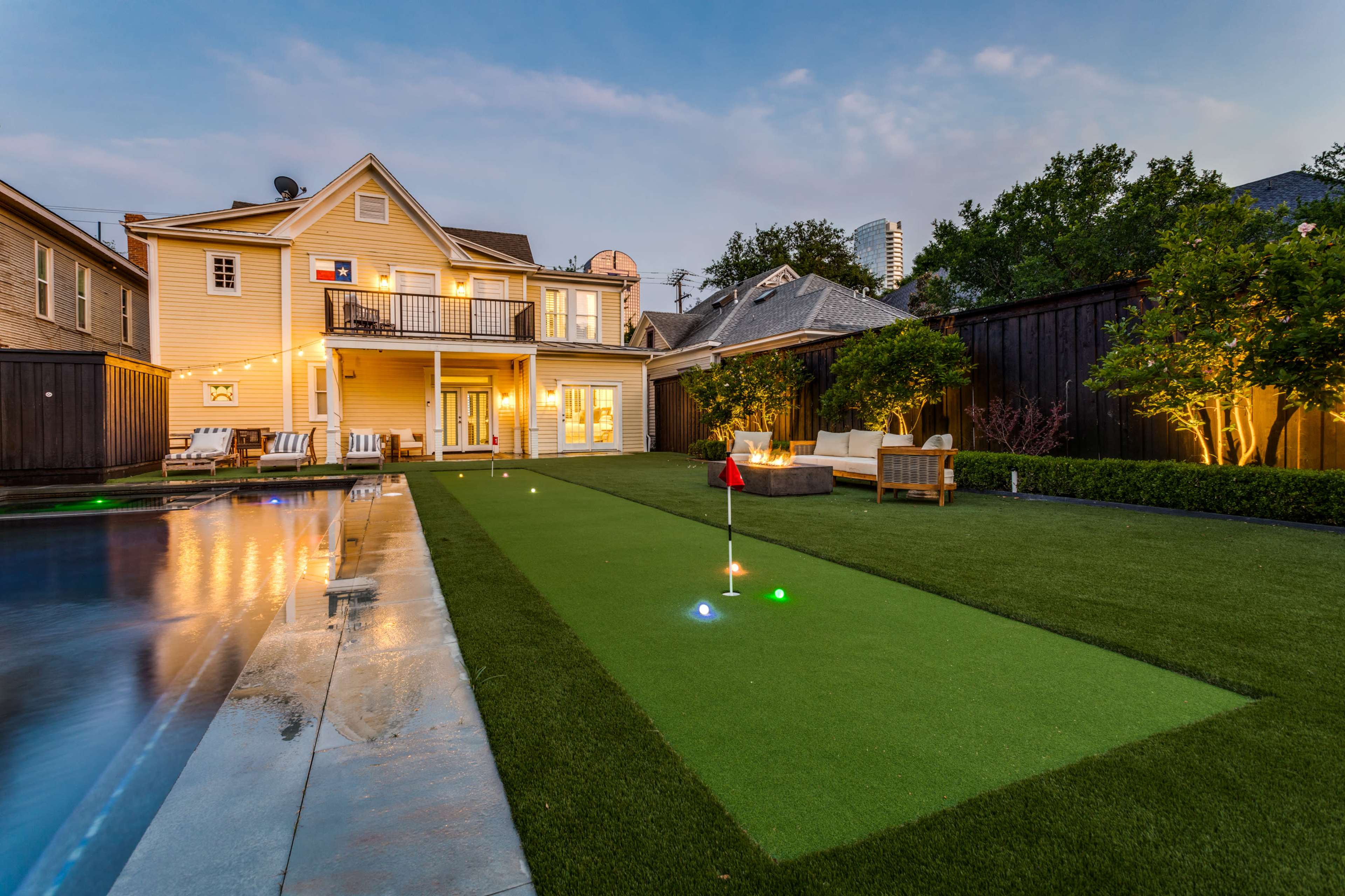 A two-story yellow house features a well-maintained backyard with a putting green and a swimming pool, illuminated by landscaping lights at dusk.