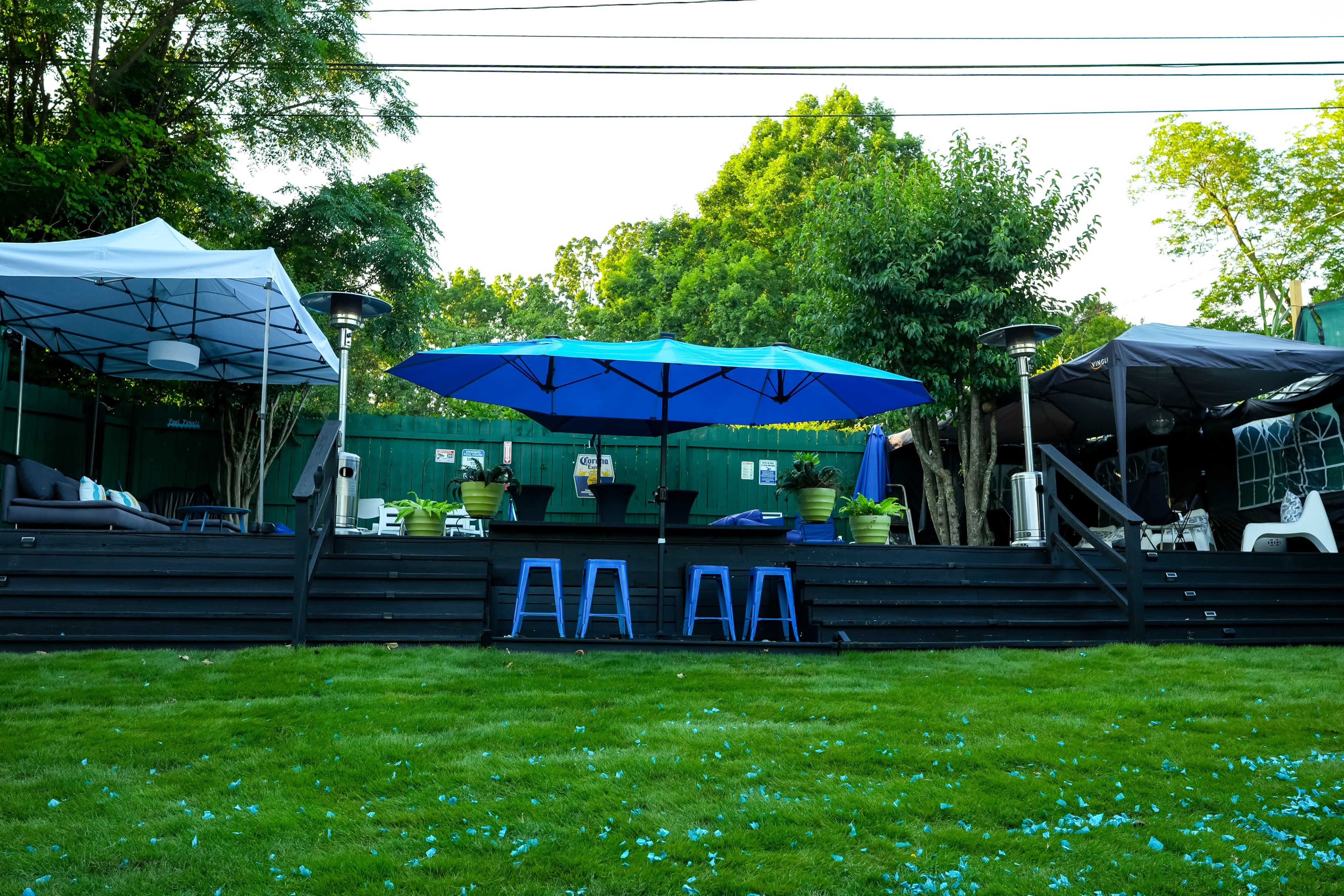 The image shows a backyard patio with two large canopies, multiple seating areas, and an umbrella over a central table surrounded by greenery.