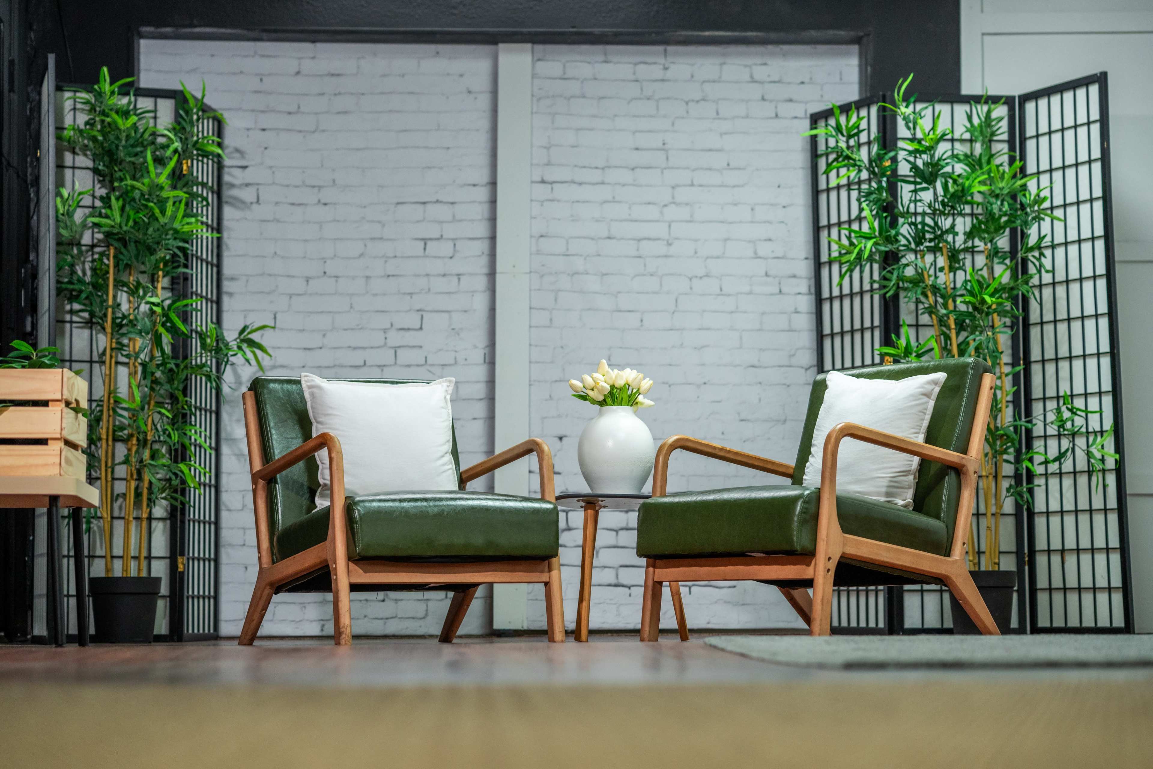 The image shows a cozy seating arrangement with two green chairs, a small round table, and decorative plants against a brick wall backdrop.