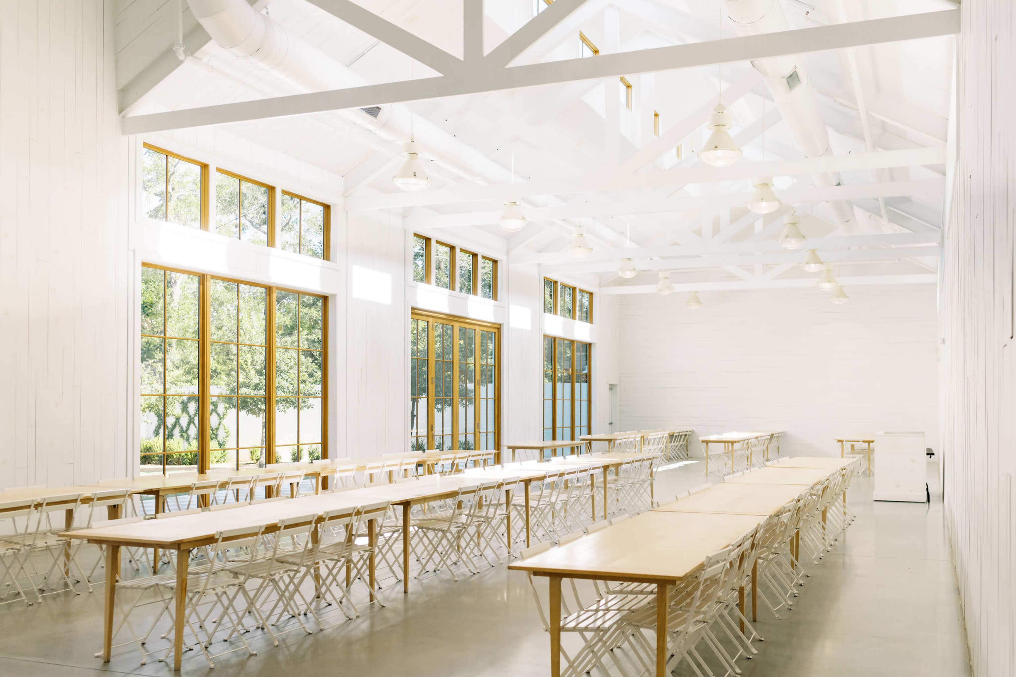 The image shows a spacious, well-lit venue with rows of long tables and large windows overlooking greenery.