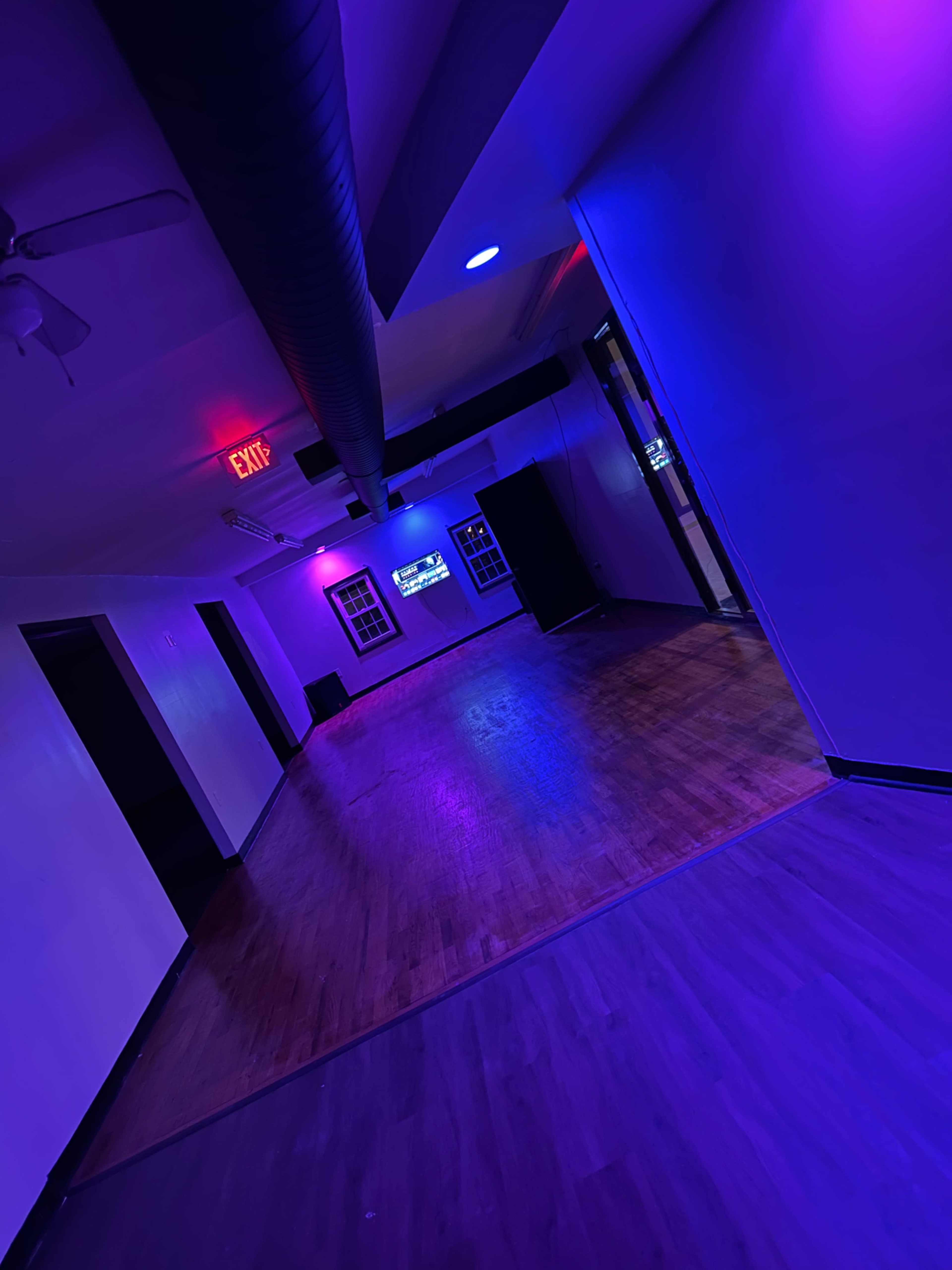 A dimly lit hallway with purple and blue lights, wooden flooring, and an exit sign in the background.