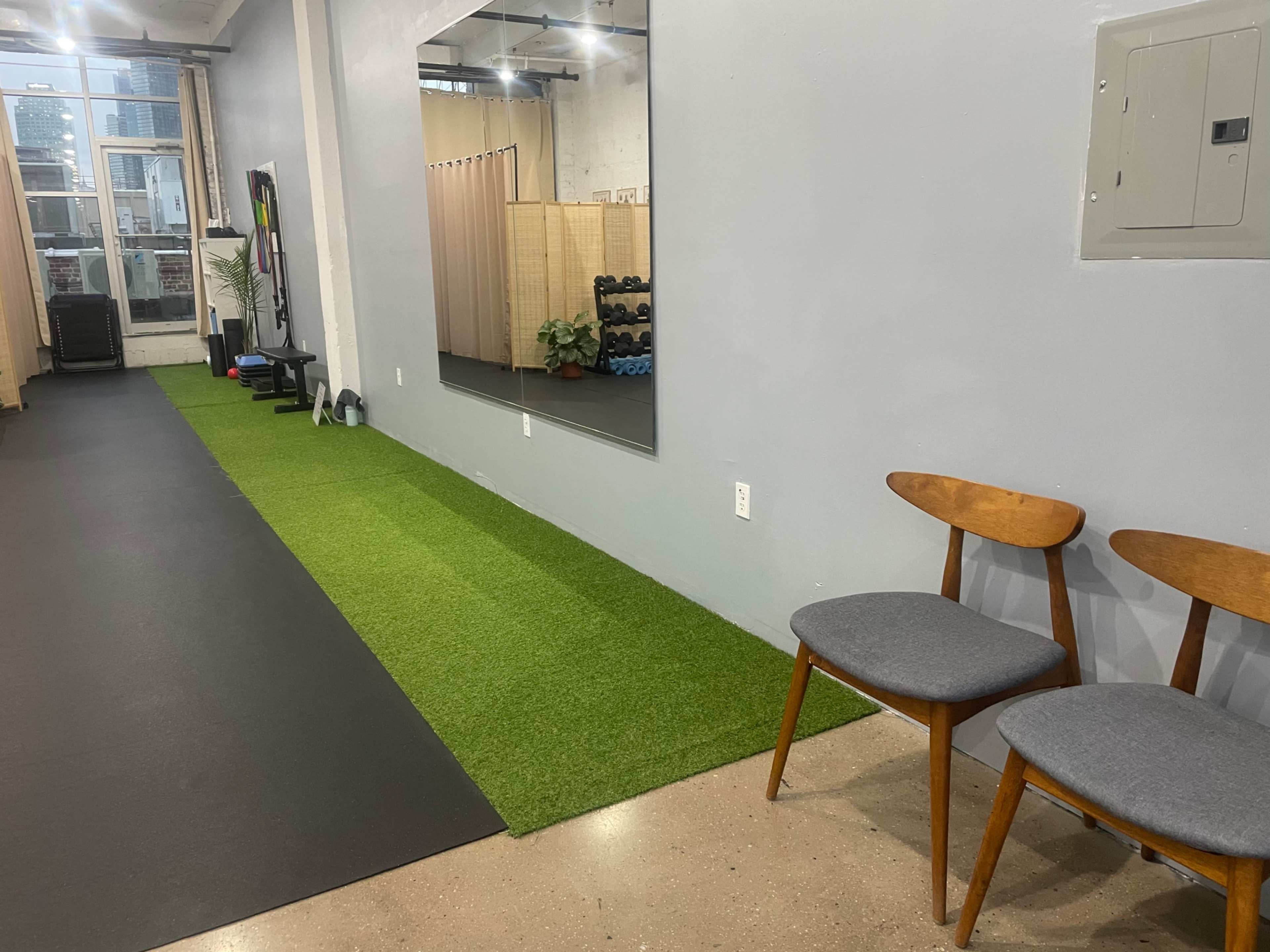 The image shows a fitness space with artificial grass flooring, workout equipment in the background, and two wooden chairs positioned nearby.