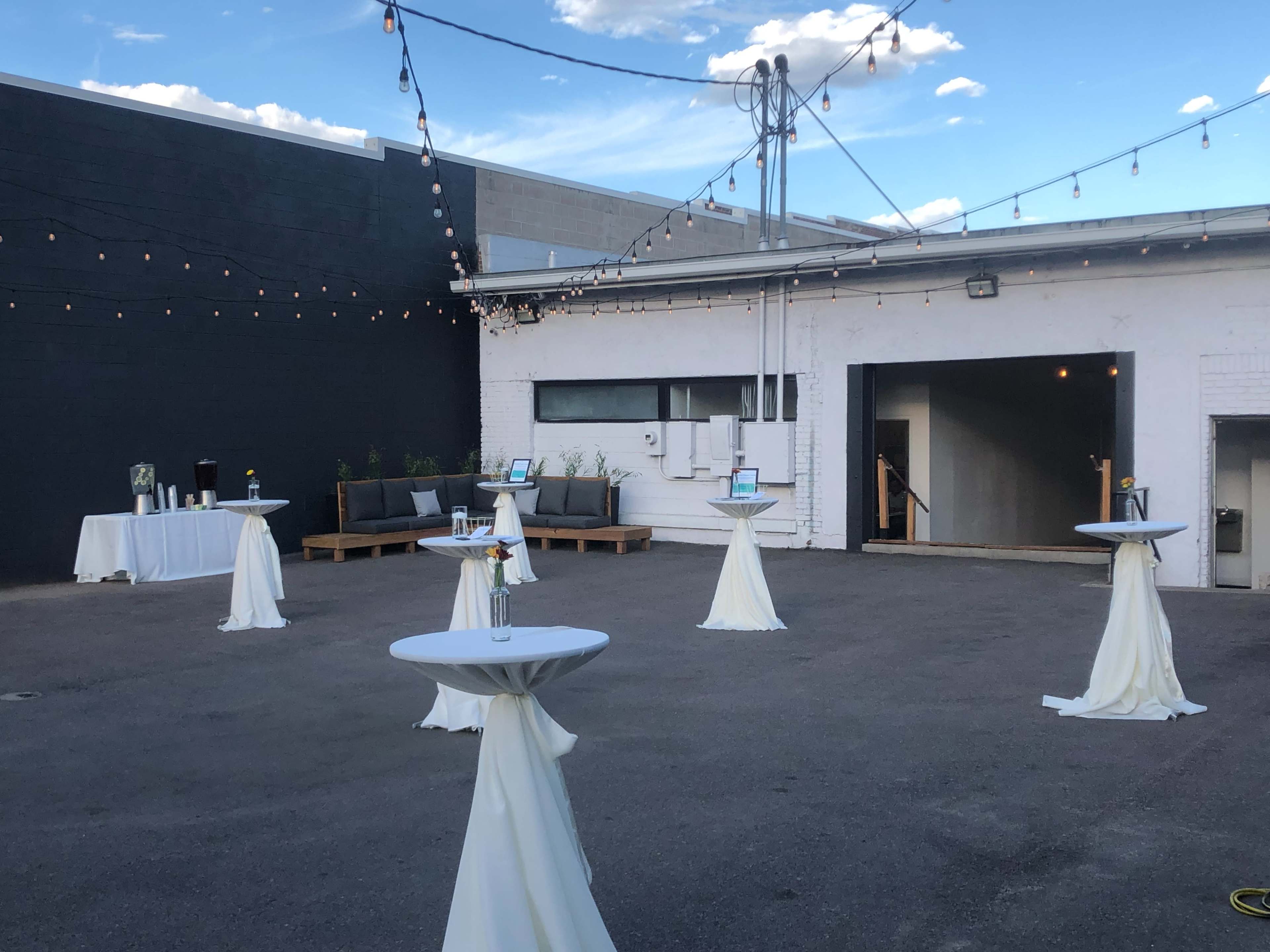 An outdoor event space features tables with white tablecloths and string lights above, ready for a gathering.