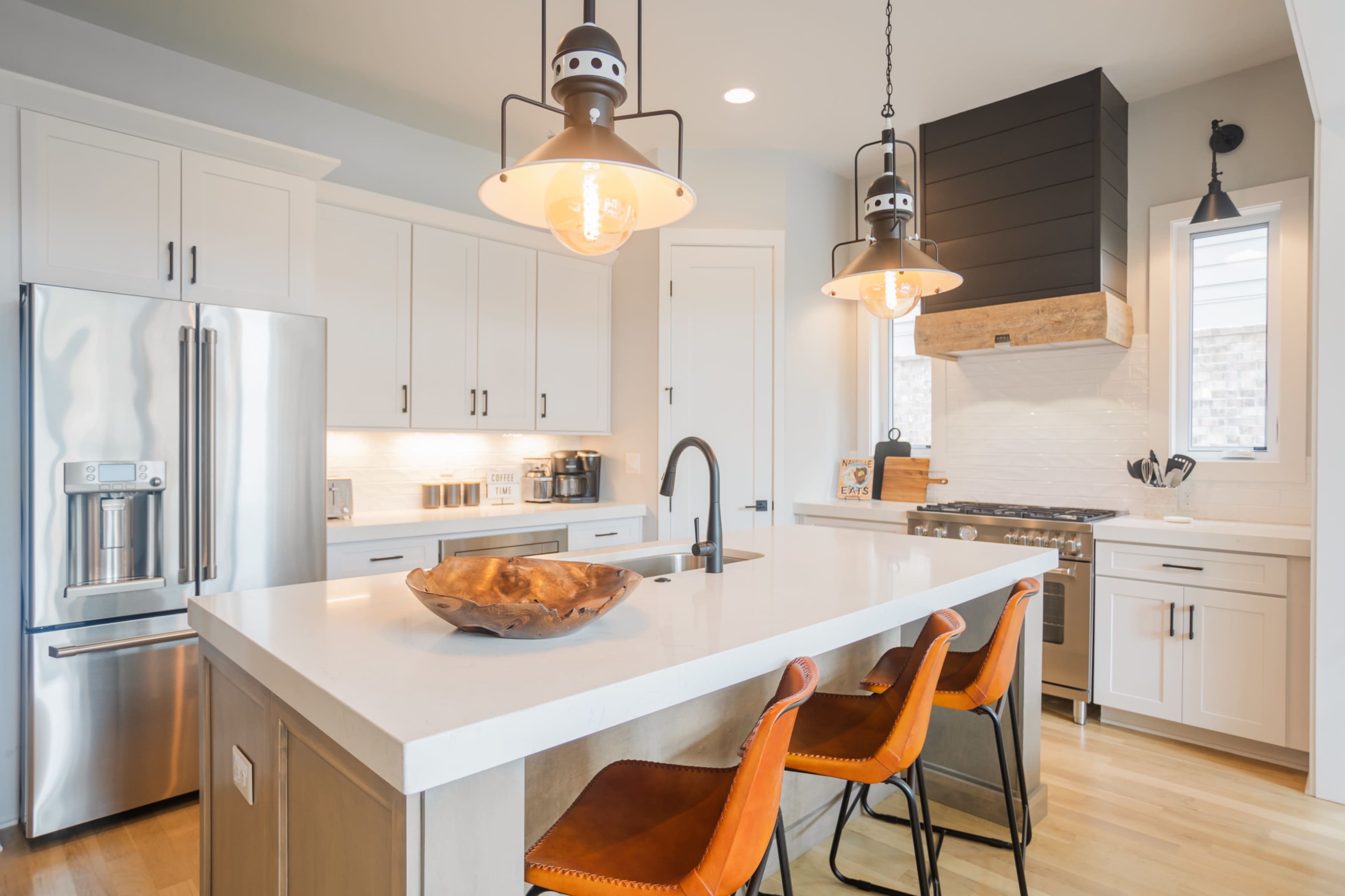 The image shows a modern kitchen featuring white cabinetry, stainless steel appliances, a large island with a stone countertop, and bar stools.