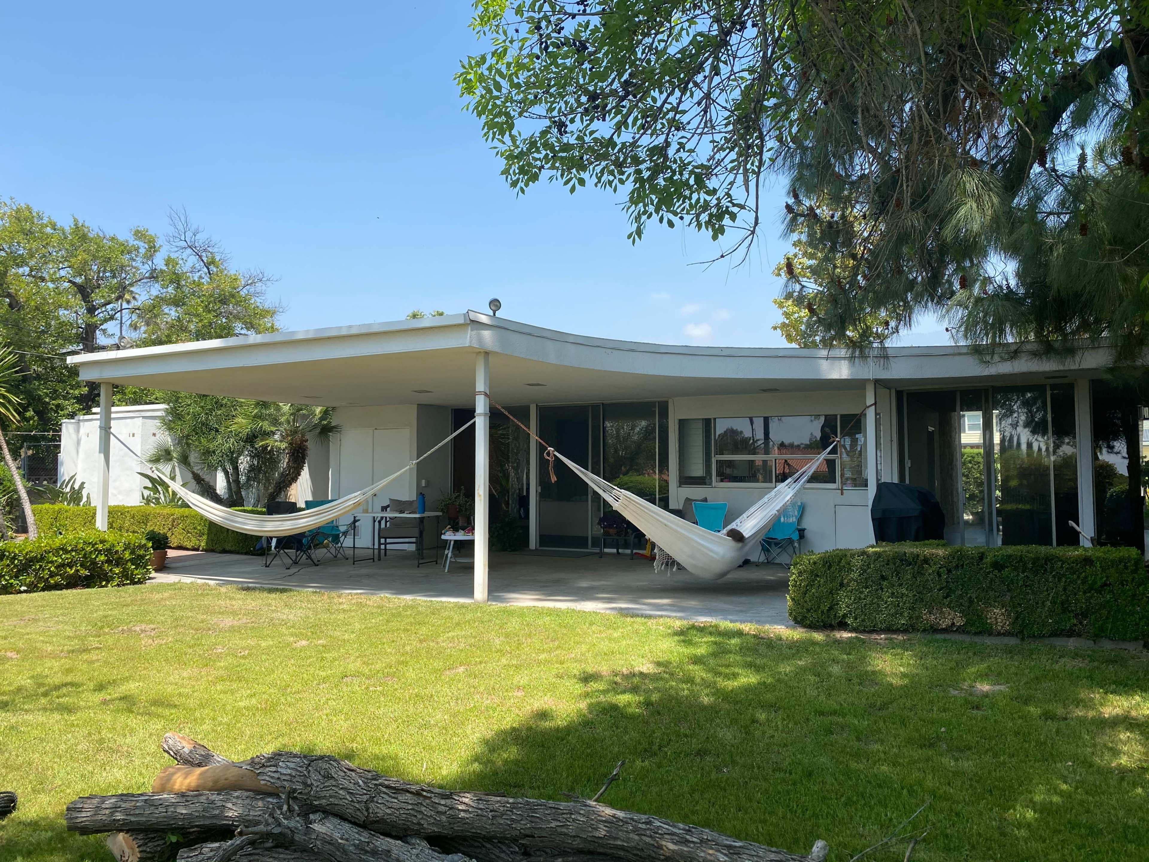 A modern house with a large, shaded patio featuring two hammocks, surrounded by a well-maintained lawn and shrubs.