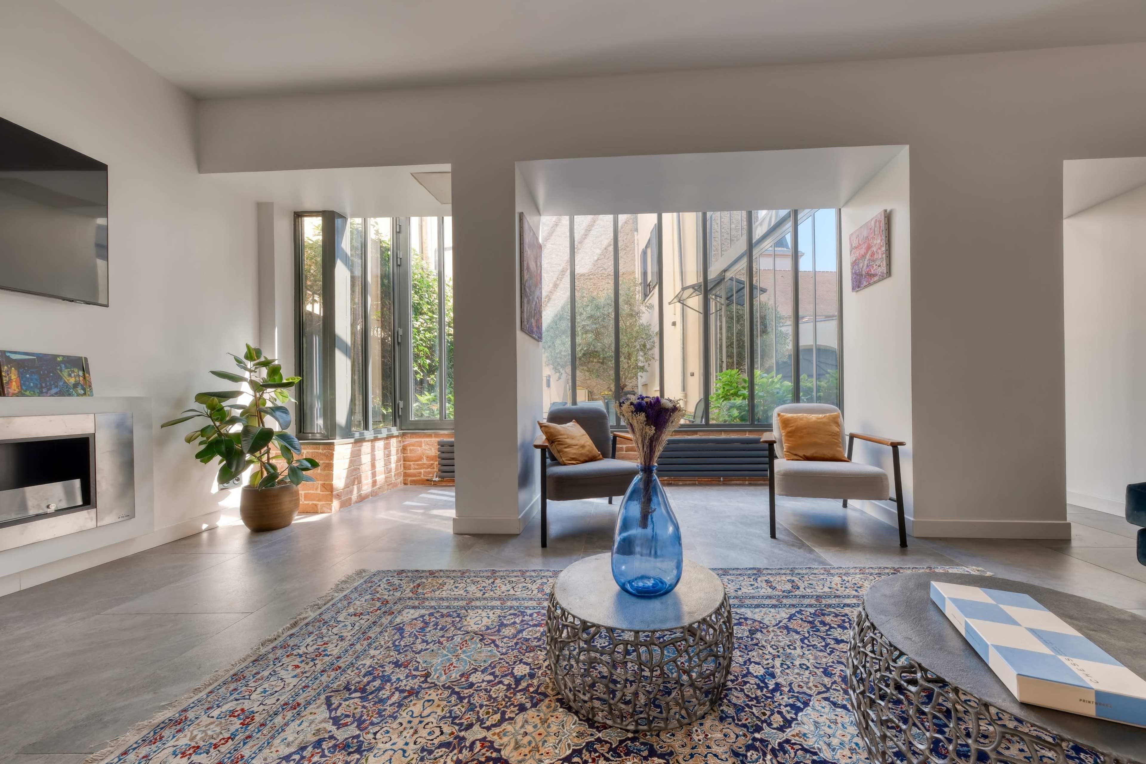 The image shows a modern interior living space with two armchairs, a patterned area rug, and a decorative table, all illuminated by natural light coming through large windows.
