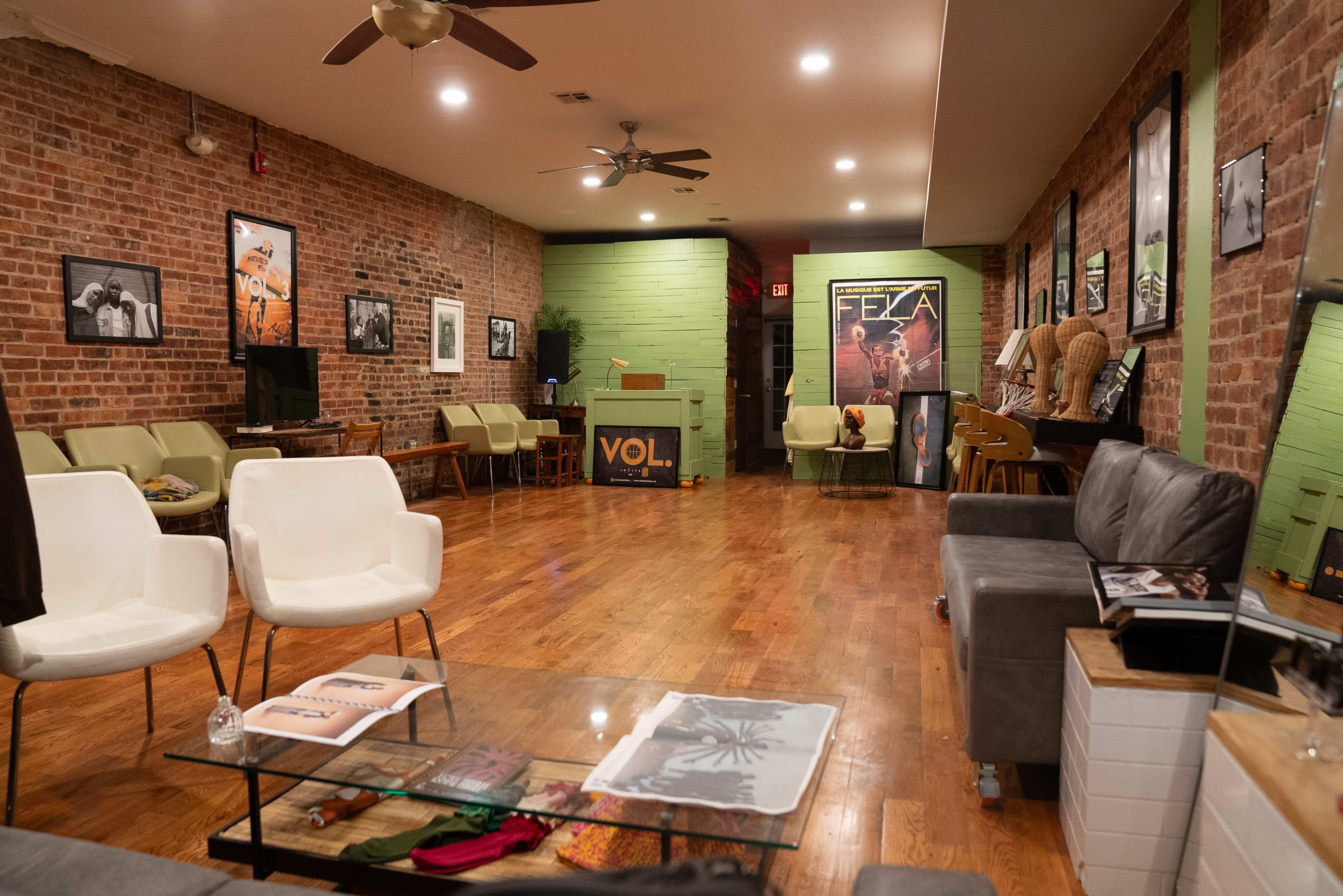 The image shows a spacious lounge area with hardwood floors, brick walls, various wall art, and seating arrangements that include white chairs and a gray sofa.