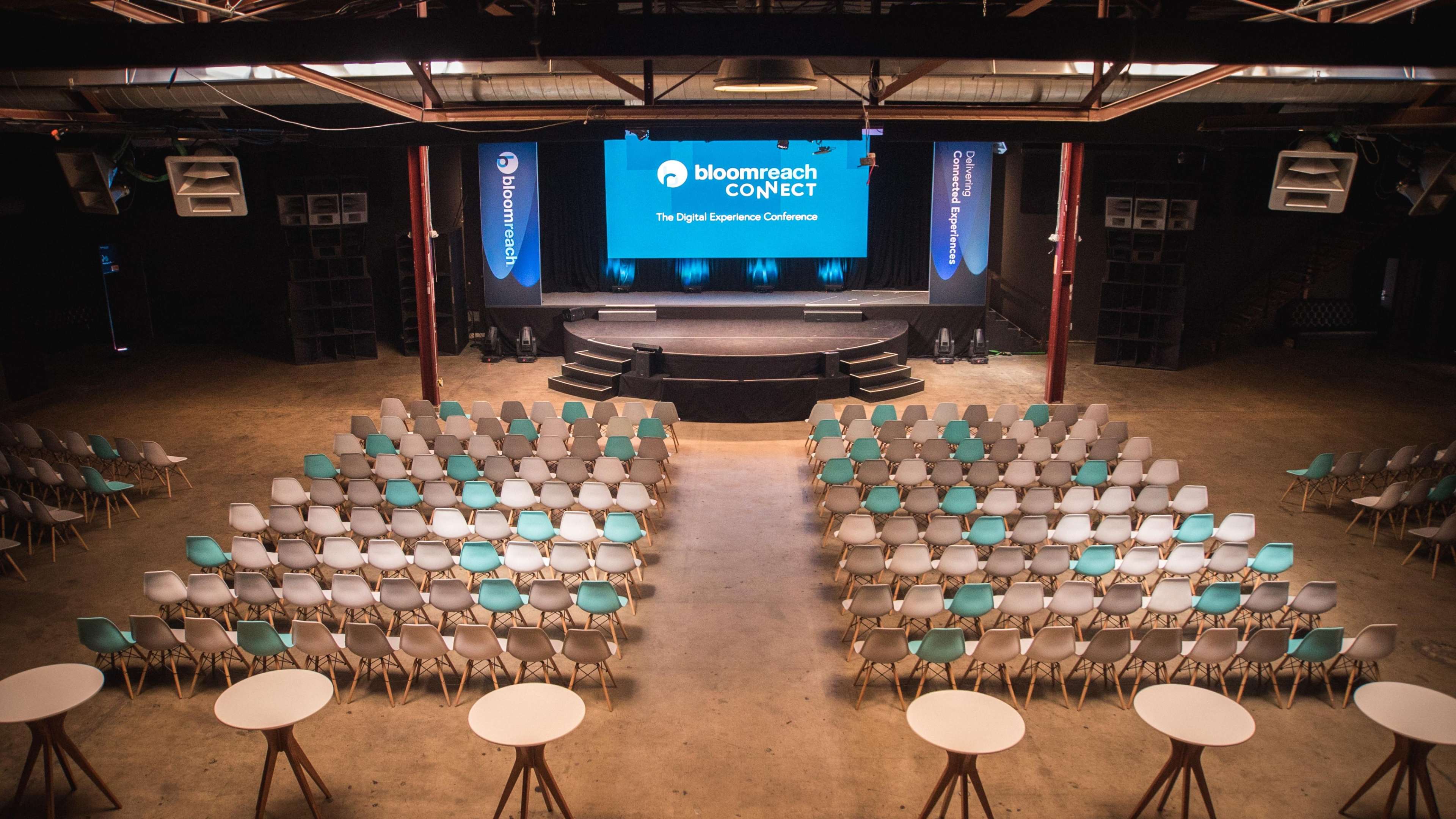 The image shows a spacious conference venue set up for the bloomreach CONNECT event, featuring rows of chairs facing a stage with a large screen.