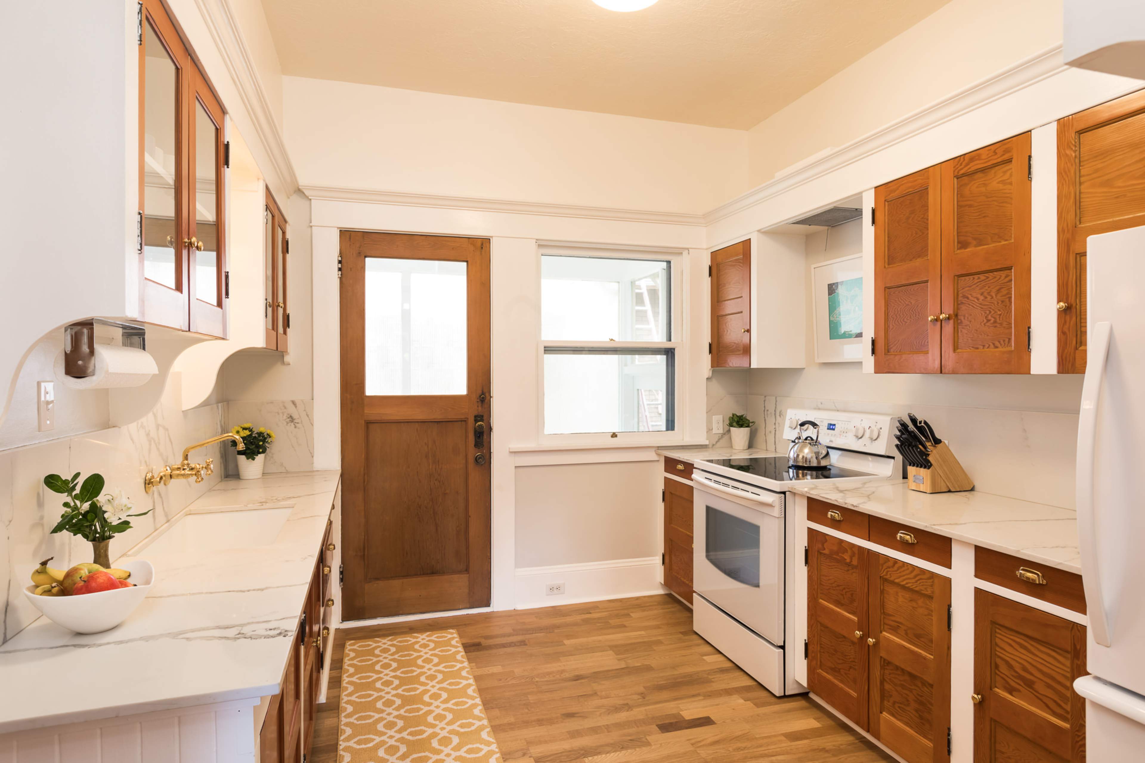 A kitchen features white walls, wooden cabinetry, and a marble countertop with a window and door leading outside.
