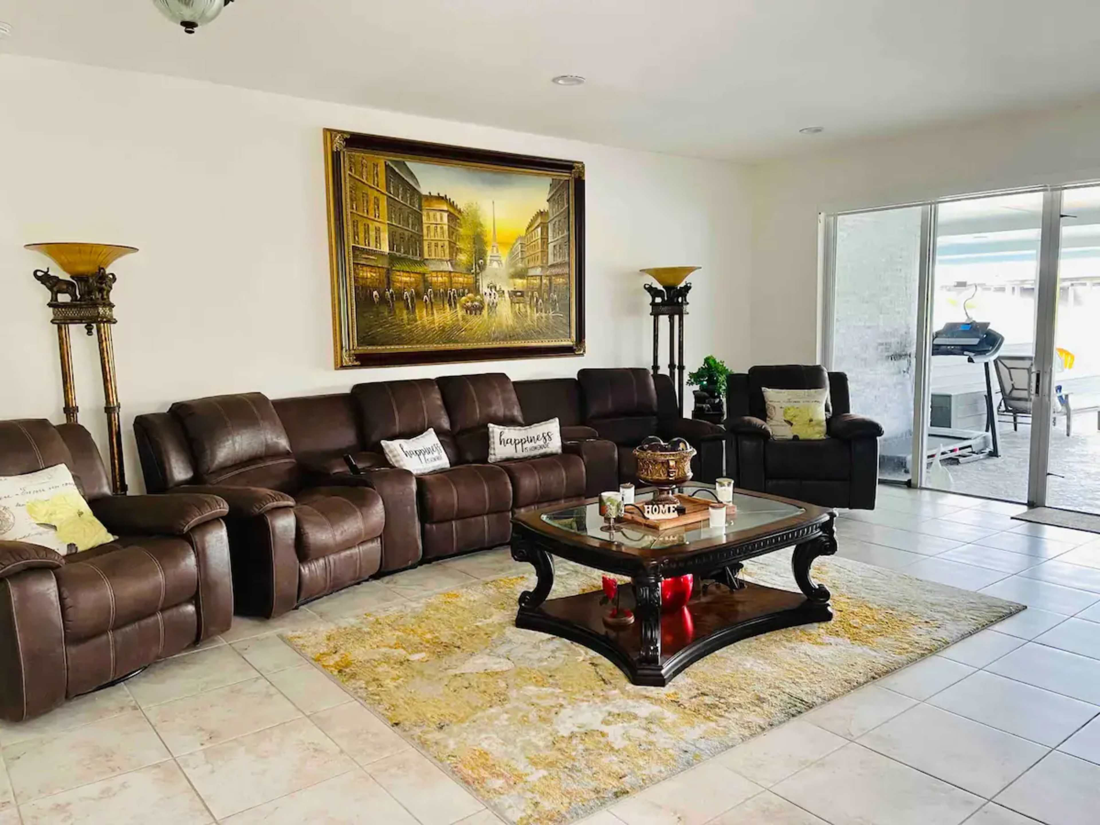 A living room with a brown leather sectional sofa, a glass coffee table, a decorative area rug, and large windows leading to an outdoor space.