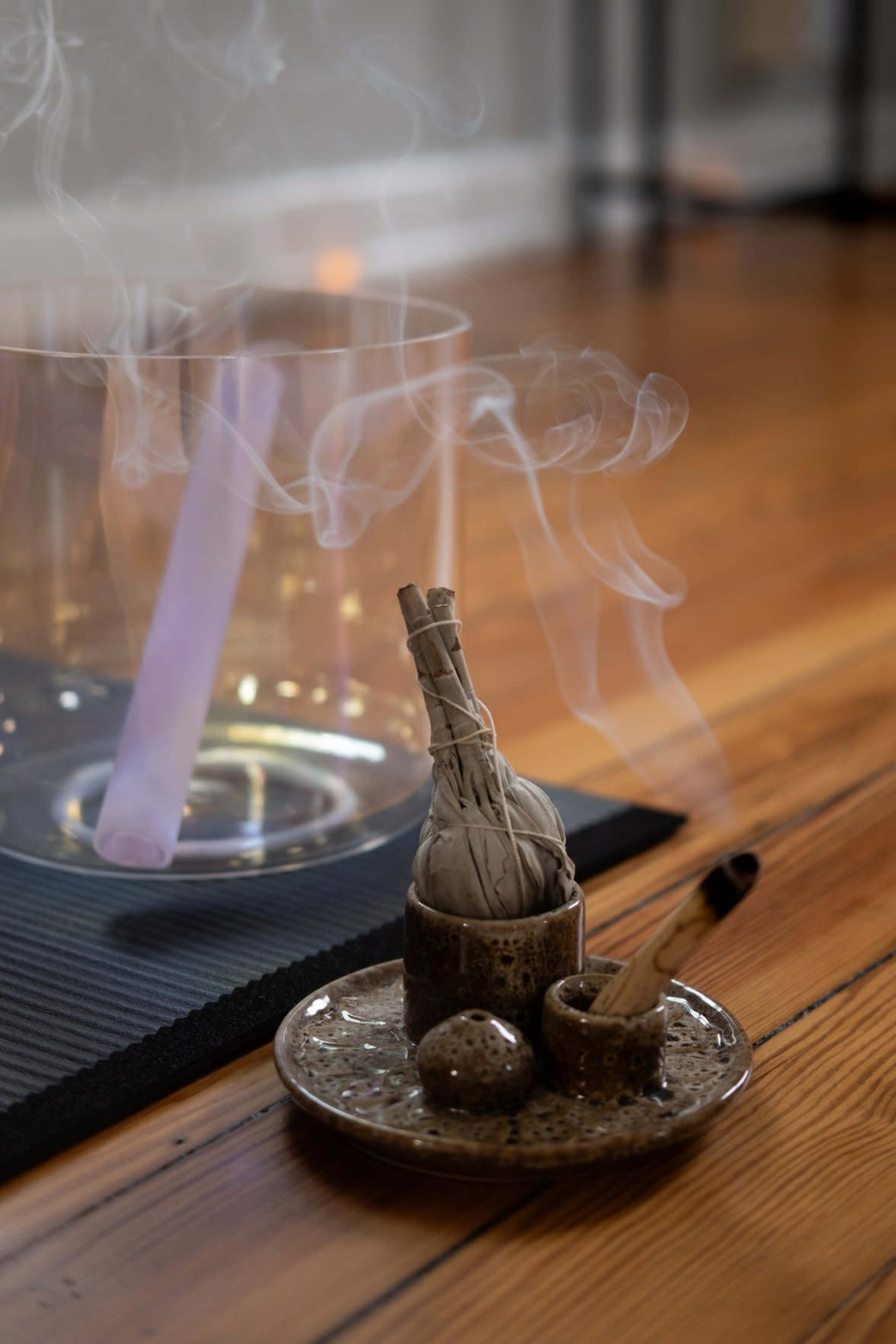 A bundle of sage is burning on a small plate beside a glass container, with wisps of smoke rising in a room featuring wooden flooring.