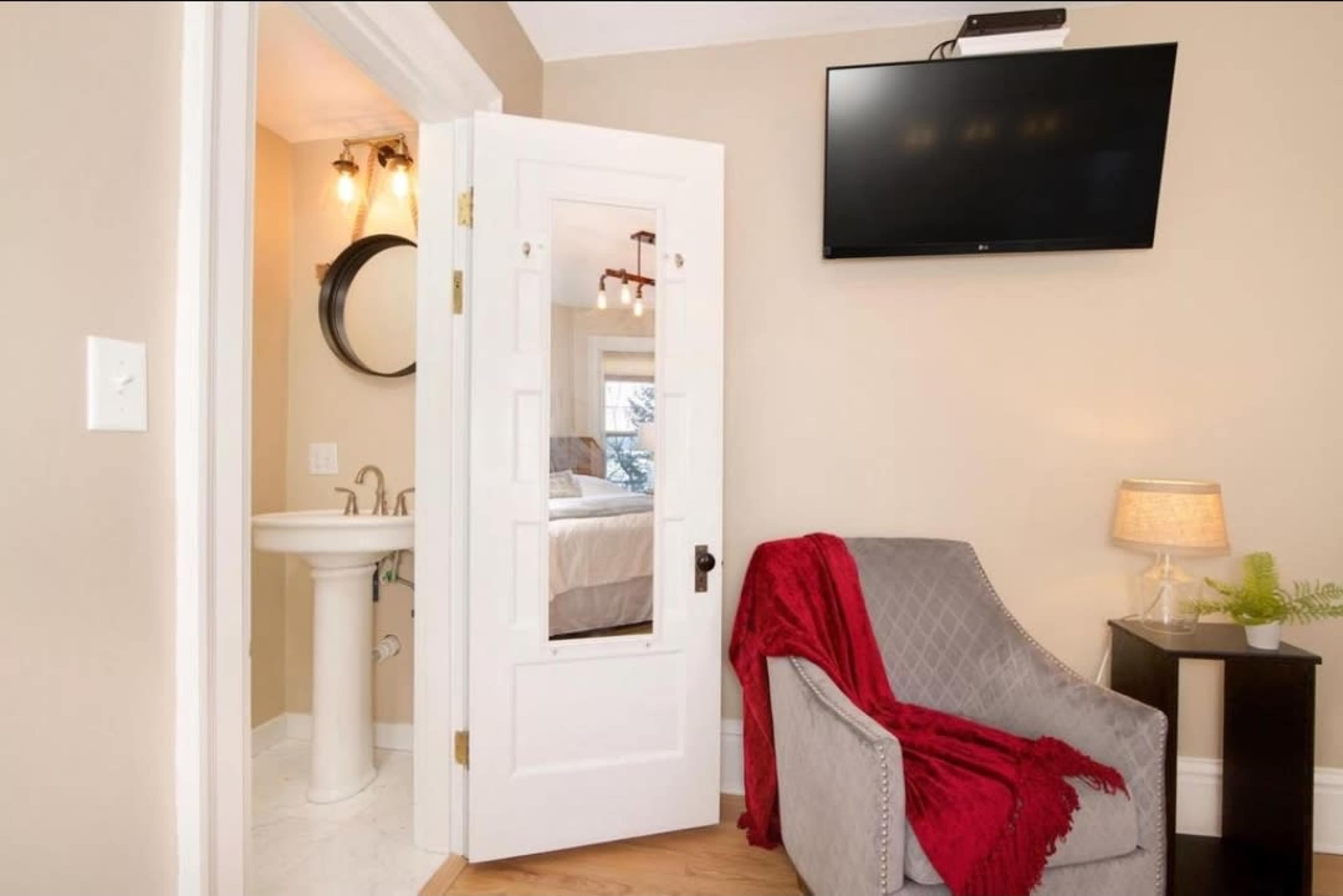 A bathroom door opens into a small room featuring a gray chair, a table with a lamp, and a wall-mounted television.