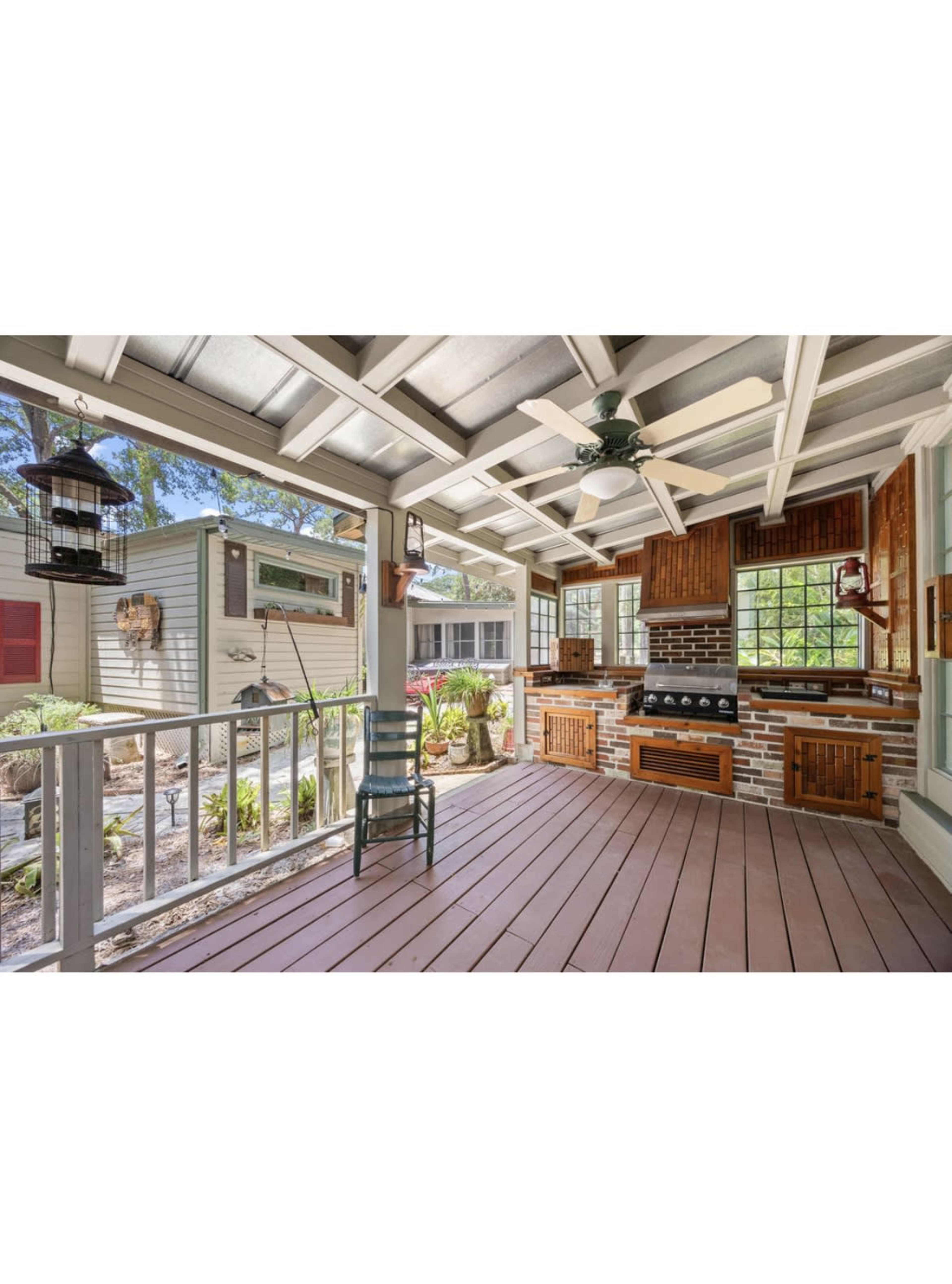 The image shows an outdoor kitchen area with a grill, wooden cabinetry, and seating, surrounded by greenery.