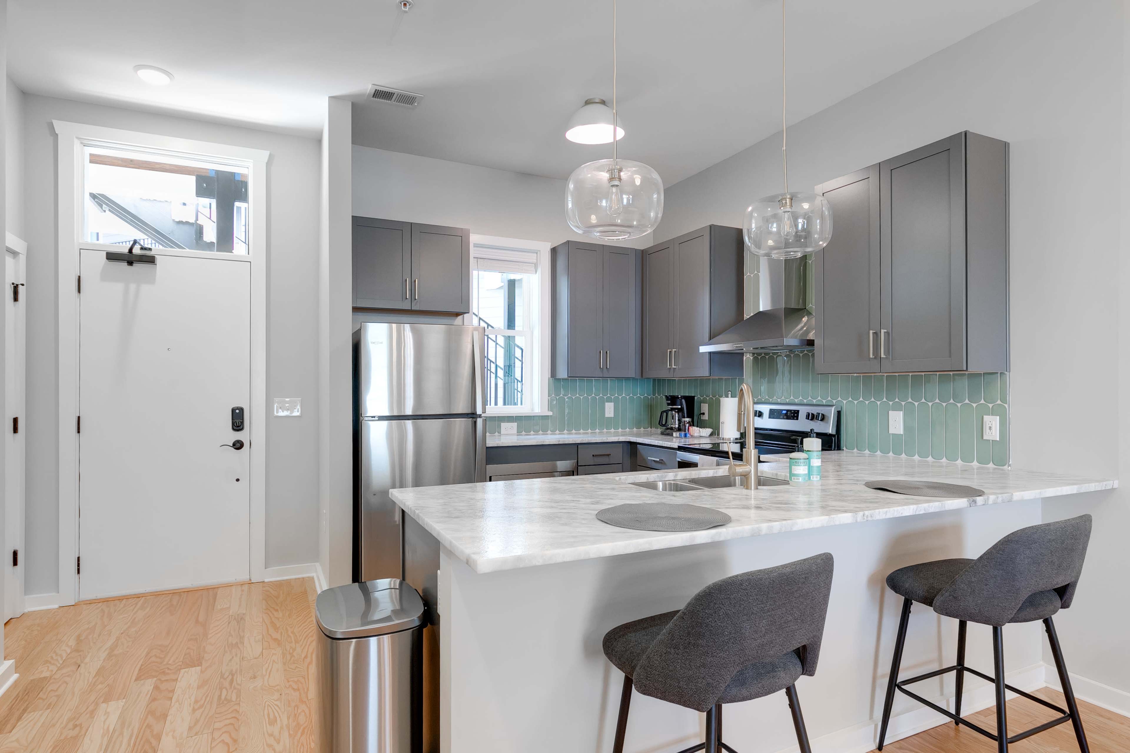 A modern kitchen with gray cabinets, a marble countertop, and two bar stools, connected to a living area with a door leading outside.