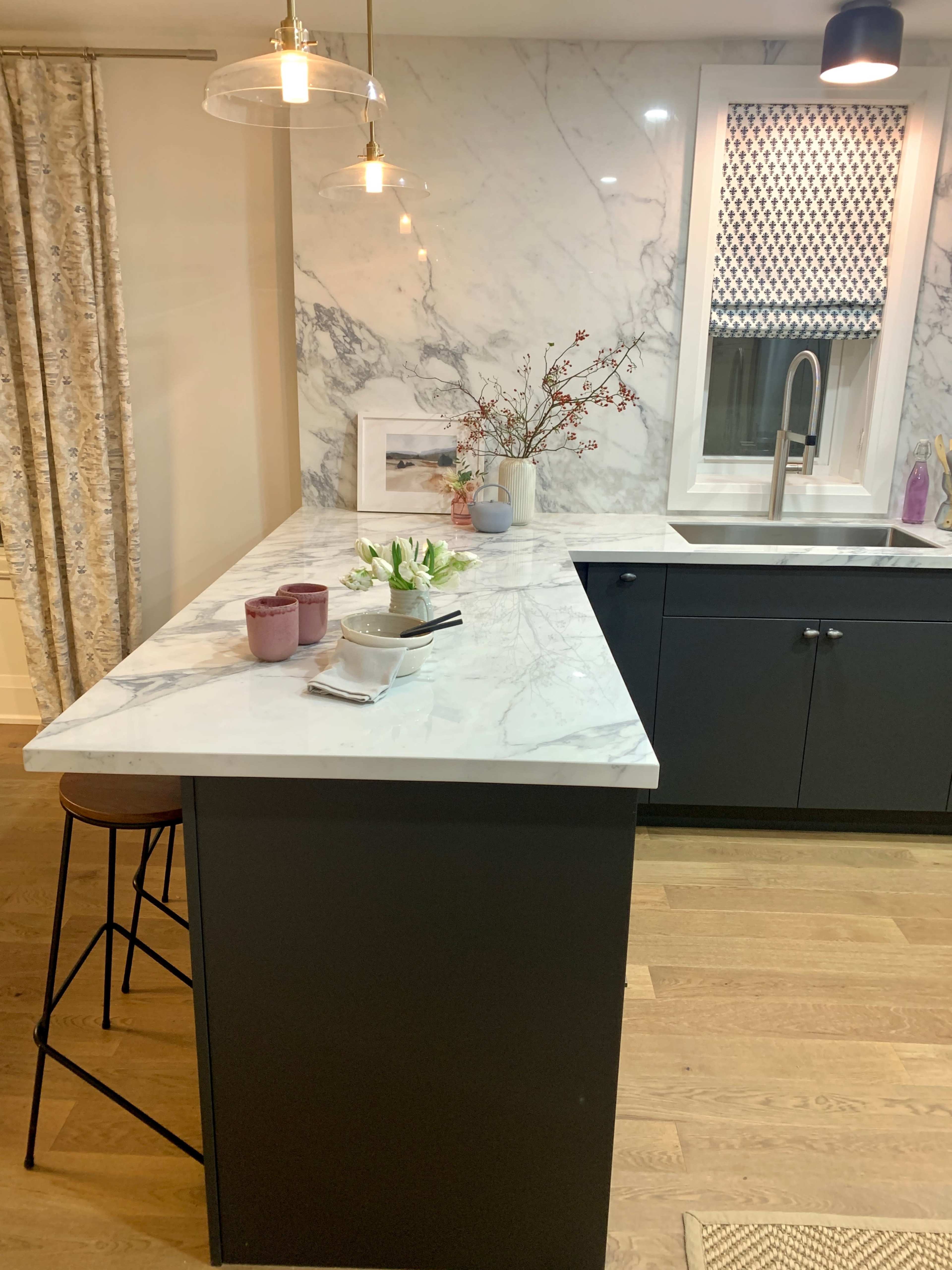 A modern kitchen featuring a marble countertop, a dark cabinetry, a sink under a window, and decorative elements on the countertop.