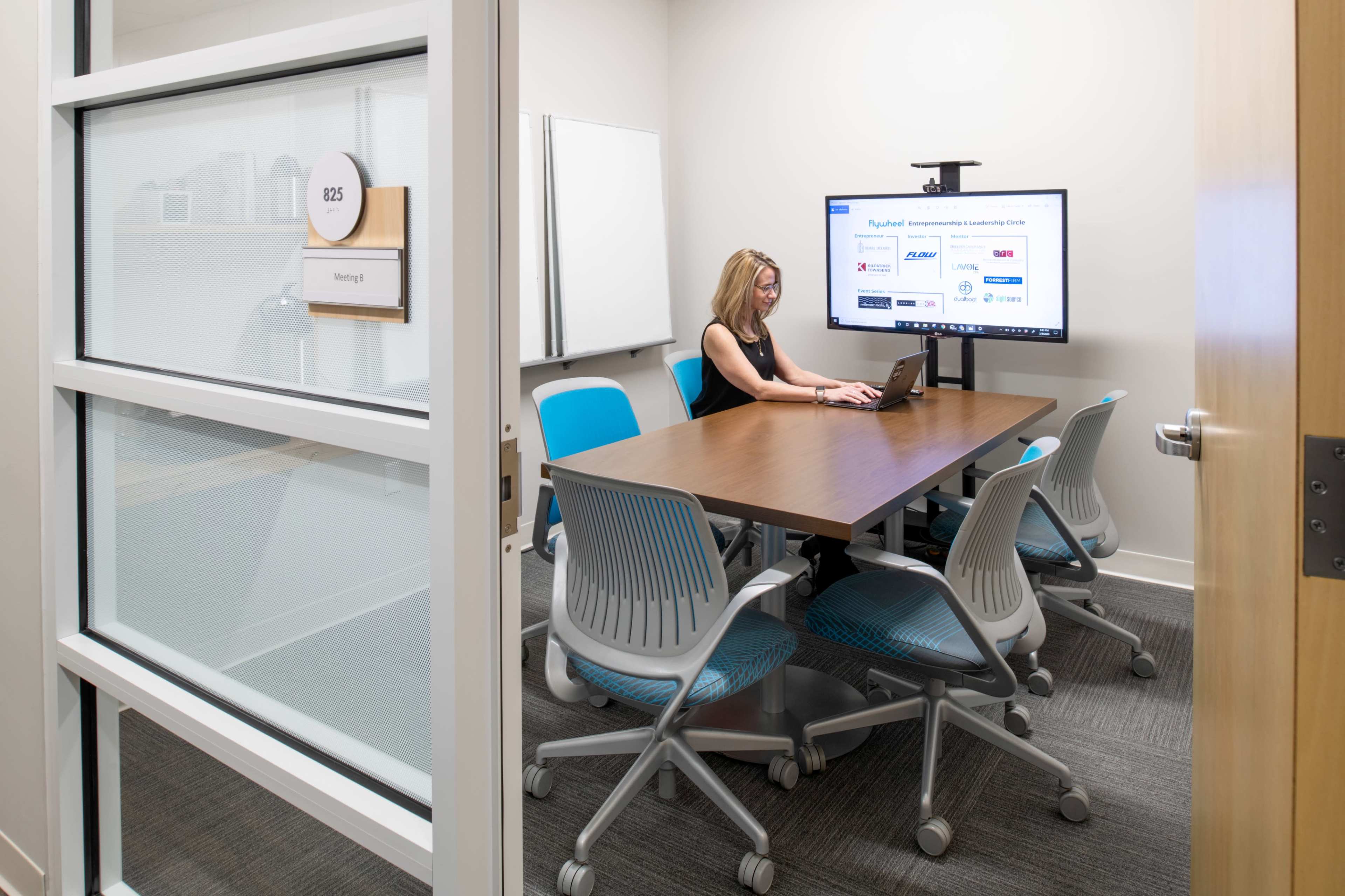 A woman sits at a conference table working on a laptop in a modern meeting room, which features a large screen displaying information.